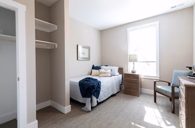 Bright single-occupancy bedroom with a twin bed, nightstand, chair and open closet by a window.