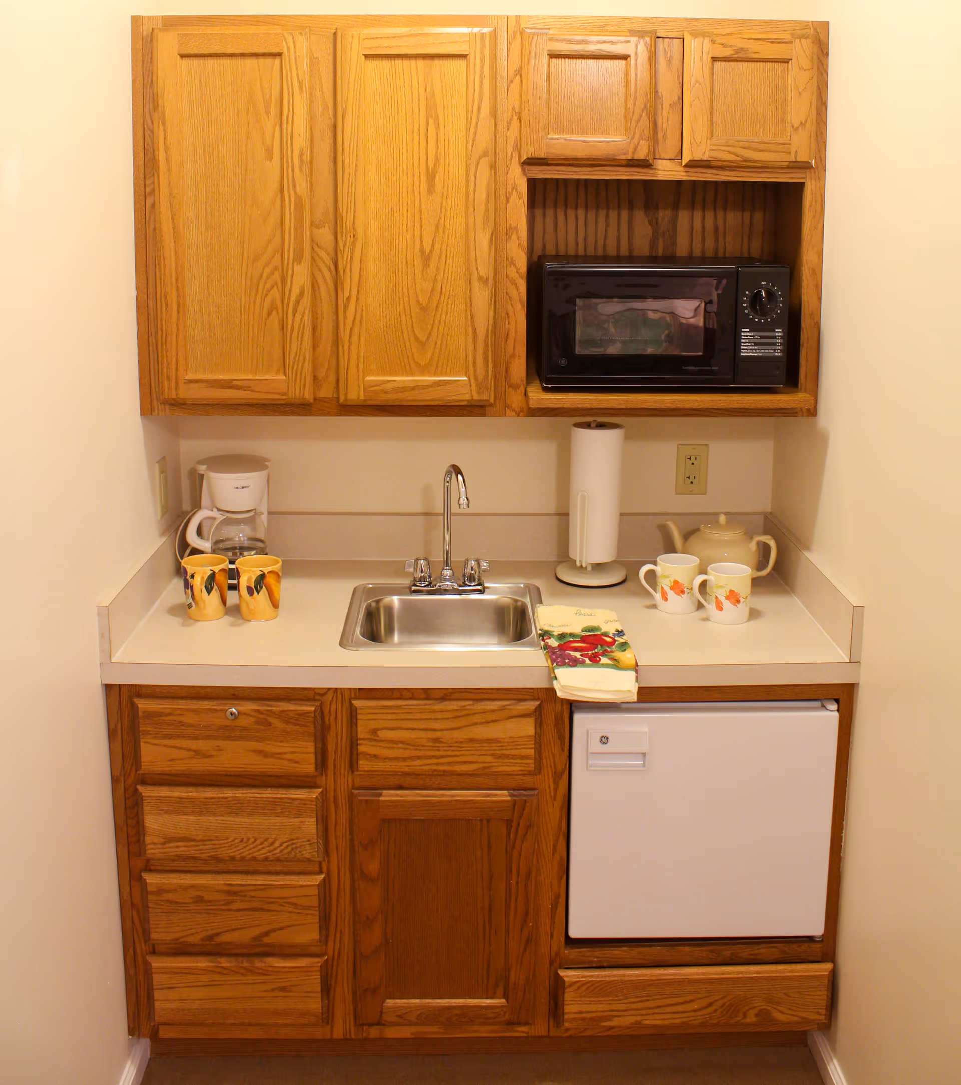 Small kitchenette with wooden cabinets, a stainless steel sink, a white mini refrigerator, a black microwave, a white coffee maker, two yellow mugs, two white mugs with autumn leaf designs, a teapot, a paper towel holder, and a folded kitchen towel on the countertop.