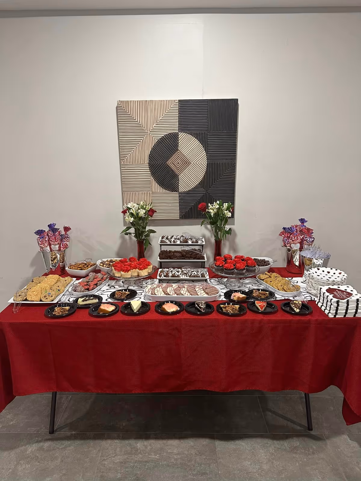 A dessert table covered with a red tablecloth displaying various sweets including cookies, cupcakes with red frosting, brownies, and other pastries. The table is decorated with two vases of flowers and two containers holding wrapped lollipops. Behind the table is a wall with a geometric black and white artwork.