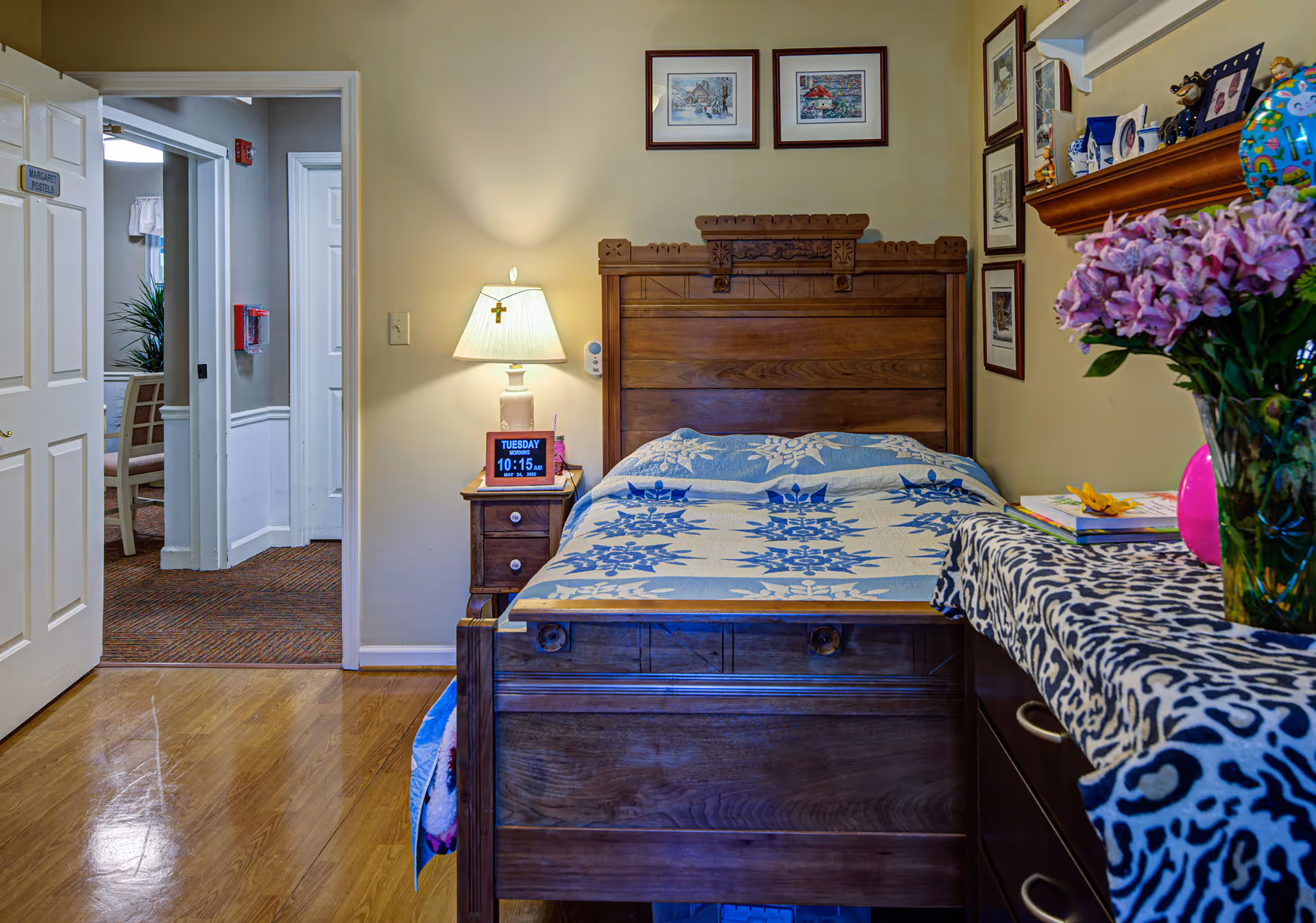 A cozy bedroom with a wooden bed featuring a blue and white patterned quilt. A nightstand with a lamp and a digital clock displaying the time and date is beside the bed. The room has framed pictures on the walls and a dresser with a leopard print cloth and a vase of purple flowers. The door is open, showing a hallway and another room with chairs and a plant.
