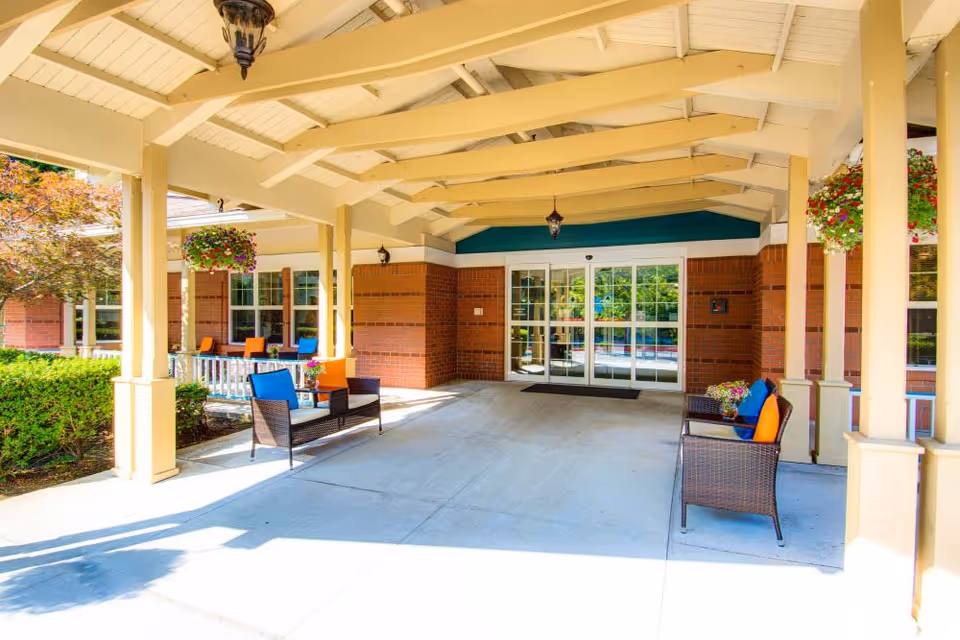 Covered entrance area of Bridgeport Place Assisted Living with brick walls, glass double doors, and outdoor seating featuring wicker chairs with colorful cushions and hanging flower baskets.