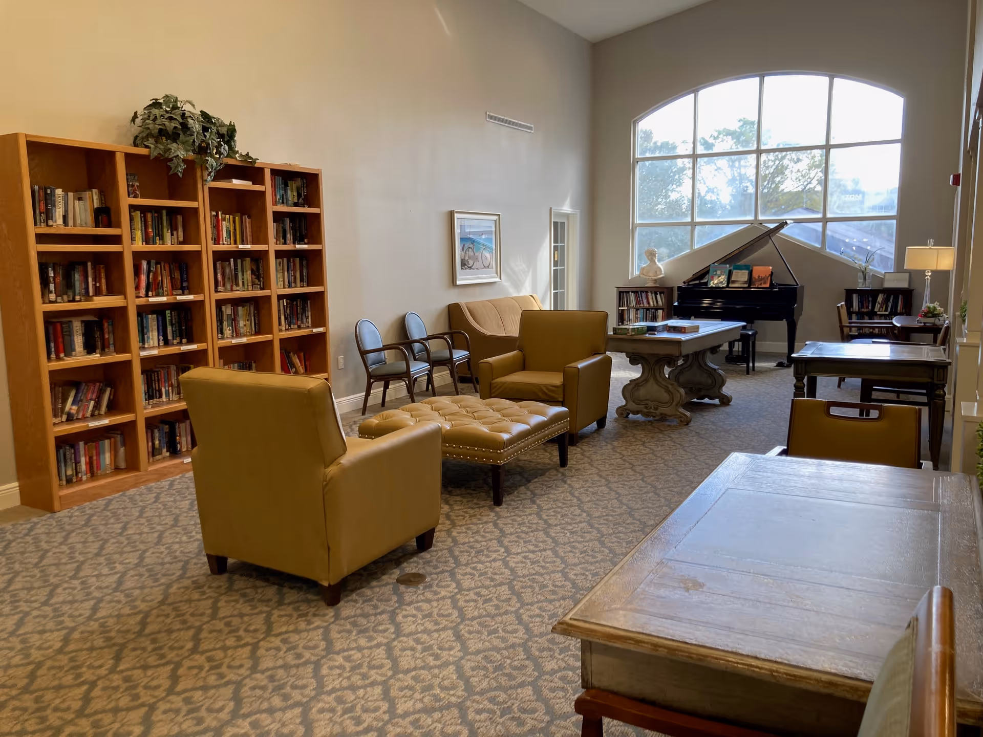 A cozy common area in a senior living facility featuring a large window letting in natural light, a grand piano, bookshelves filled with books, comfortable armchairs, a cushioned ottoman, and several tables with chairs arranged for reading or socializing.