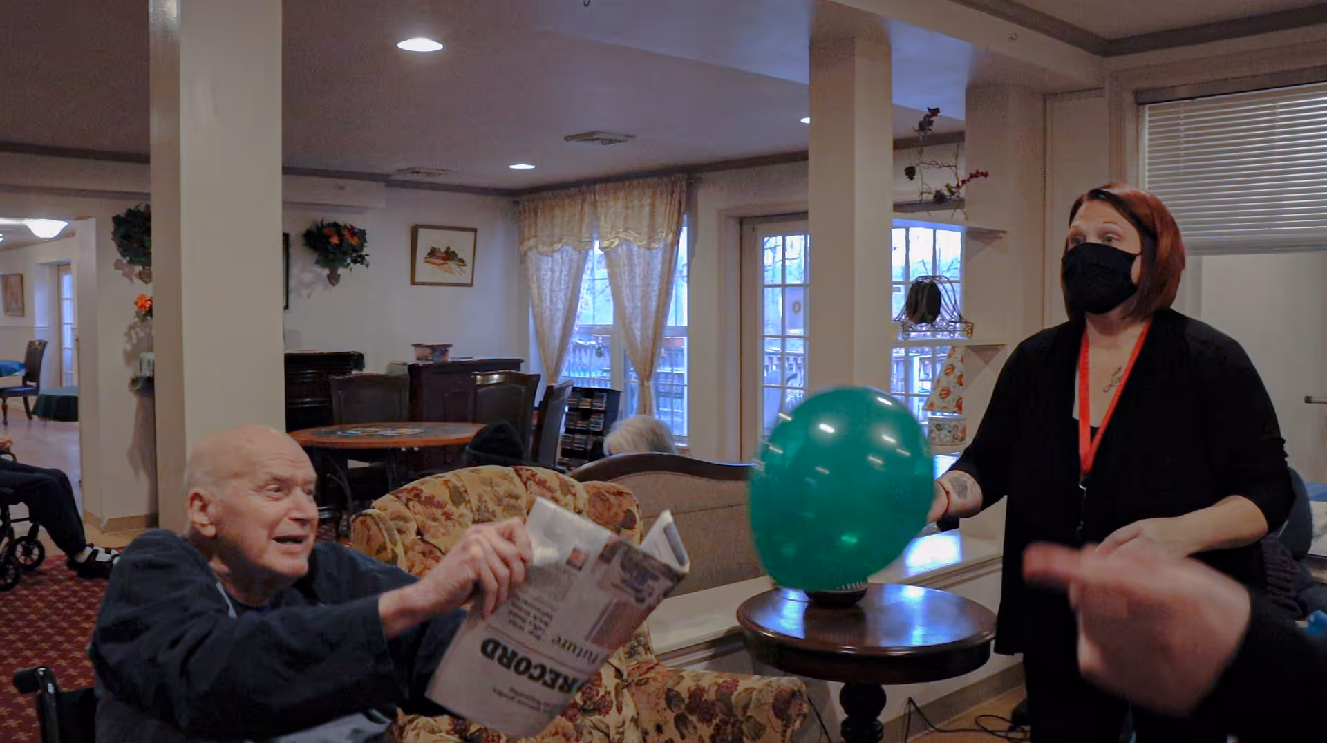 An elderly man sitting in a wheelchair holding a newspaper and reaching out towards a green balloon held by a woman wearing a black face mask and black clothing in a cozy living room setting with floral furniture and large windows with curtains.