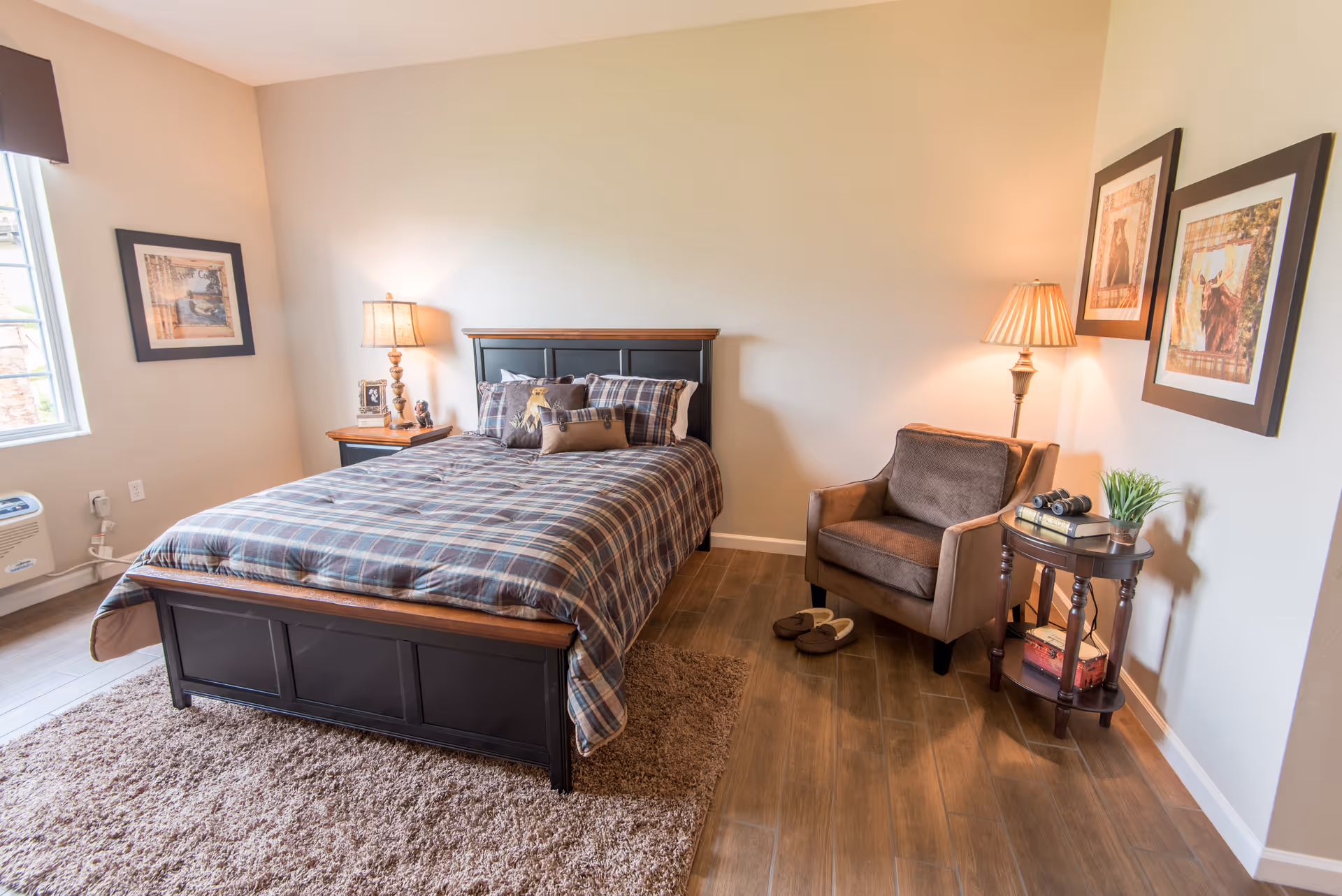 A cozy bedroom with a black wooden bed frame and plaid bedding. There is a nightstand with a lamp and decorative items on the left side of the bed. On the right side, there is a brown armchair next to a round wooden side table with a lamp, books, binoculars, and a small plant. Two framed pictures hang on the wall above the armchair. The room has wooden flooring and a beige rug partially under the bed. A window with a dark valance is visible on the left wall.