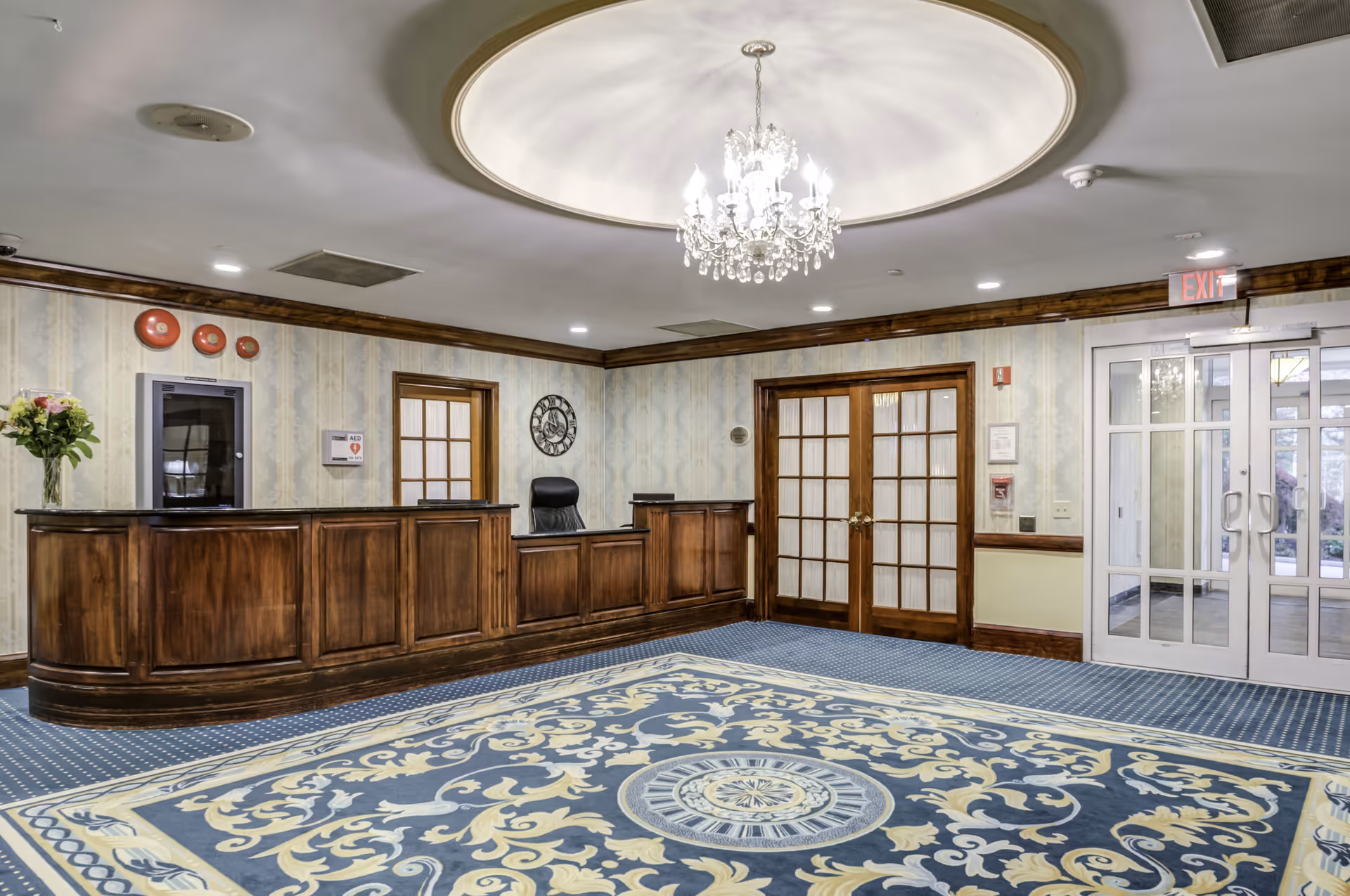 Reception area with a wooden front desk, a black office chair behind it, a large ornate blue and yellow patterned carpet on the floor, a crystal chandelier hanging from a circular recessed ceiling, floral wallpaper, double wooden doors with glass panels, and glass exit doors leading outside.