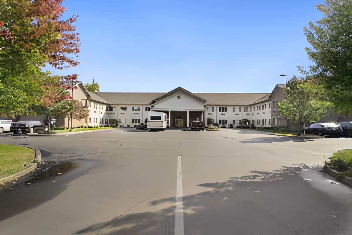 Front view of a two-story senior living facility with a covered main entrance, wide driveway/parking area, and trees on either side.