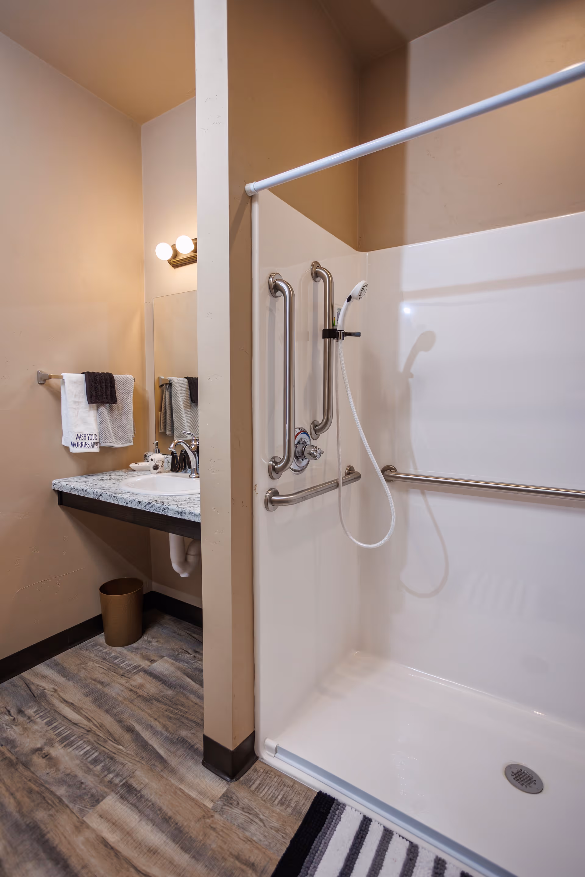 Accessible bathroom with a roll-in shower, multiple grab bars, handheld shower head and a sink with countertop and towels.