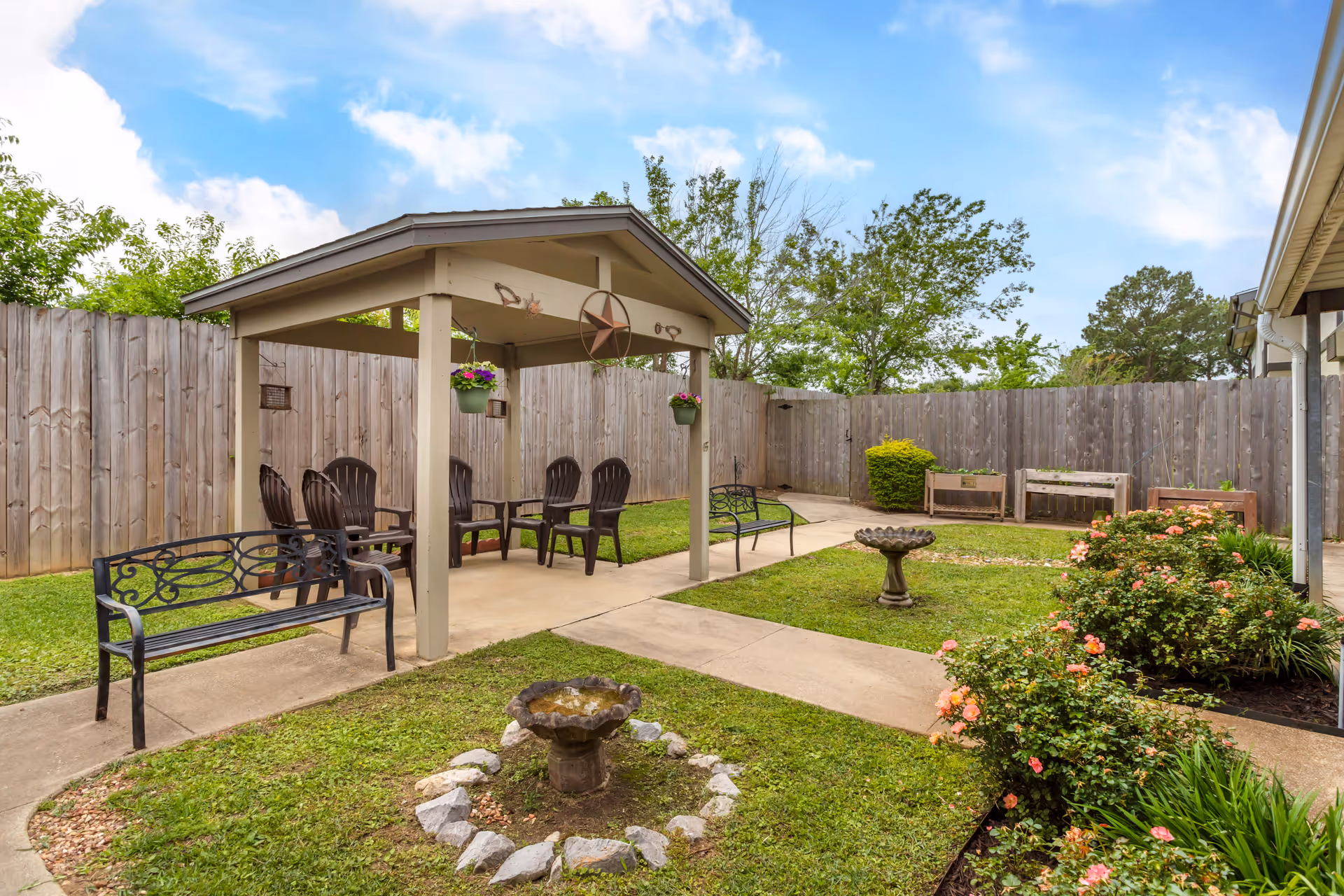 Fenced courtyard with a covered seating pavilion, benches, flowerbeds and birdbaths under a partly cloudy sky.