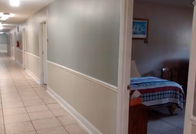 A long, tiled hallway in an assisted living facility with beige walls and white trim. Several closed doors line the hallway, and one door is open revealing a bedroom with a bed covered in a striped quilt and a bedside table with a lamp. The hallway is well-lit with ceiling lights.