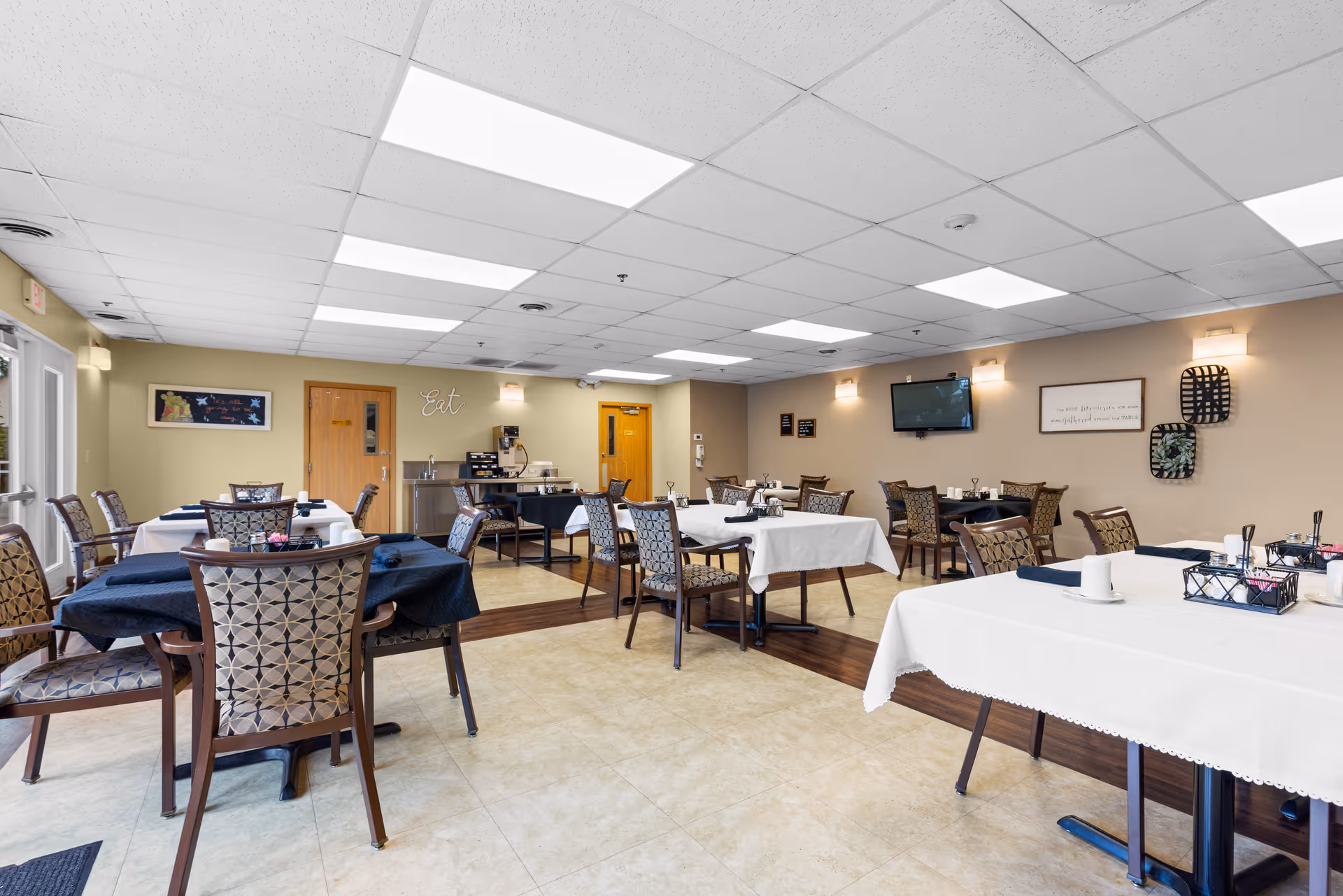 A bright dining room in an assisted living facility with multiple tables covered in white and dark blue tablecloths, surrounded by patterned chairs. The room has a tiled floor, beige walls with wall decorations, a mounted flat-screen TV, and a coffee station in the back. Large windows on the left side allow natural light to enter.