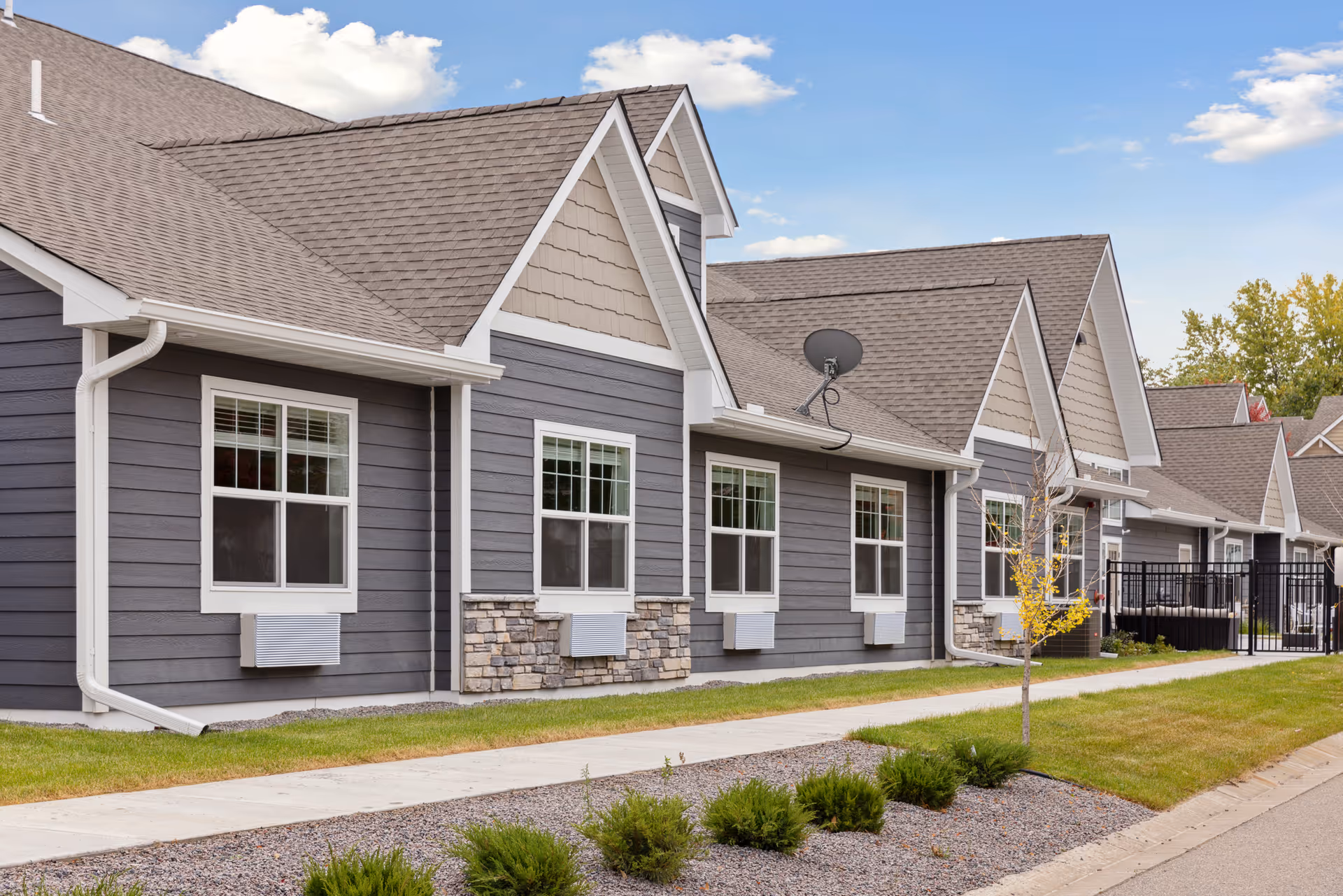 Exterior view of a senior living facility building with gray siding, white trim, multiple windows, and a stone accent on the lower part of the walls. The building has a sloped roof with shingles and a satellite dish mounted on it. There is a sidewalk and landscaped area with small bushes and a young tree in front of the building under a partly cloudy sky.