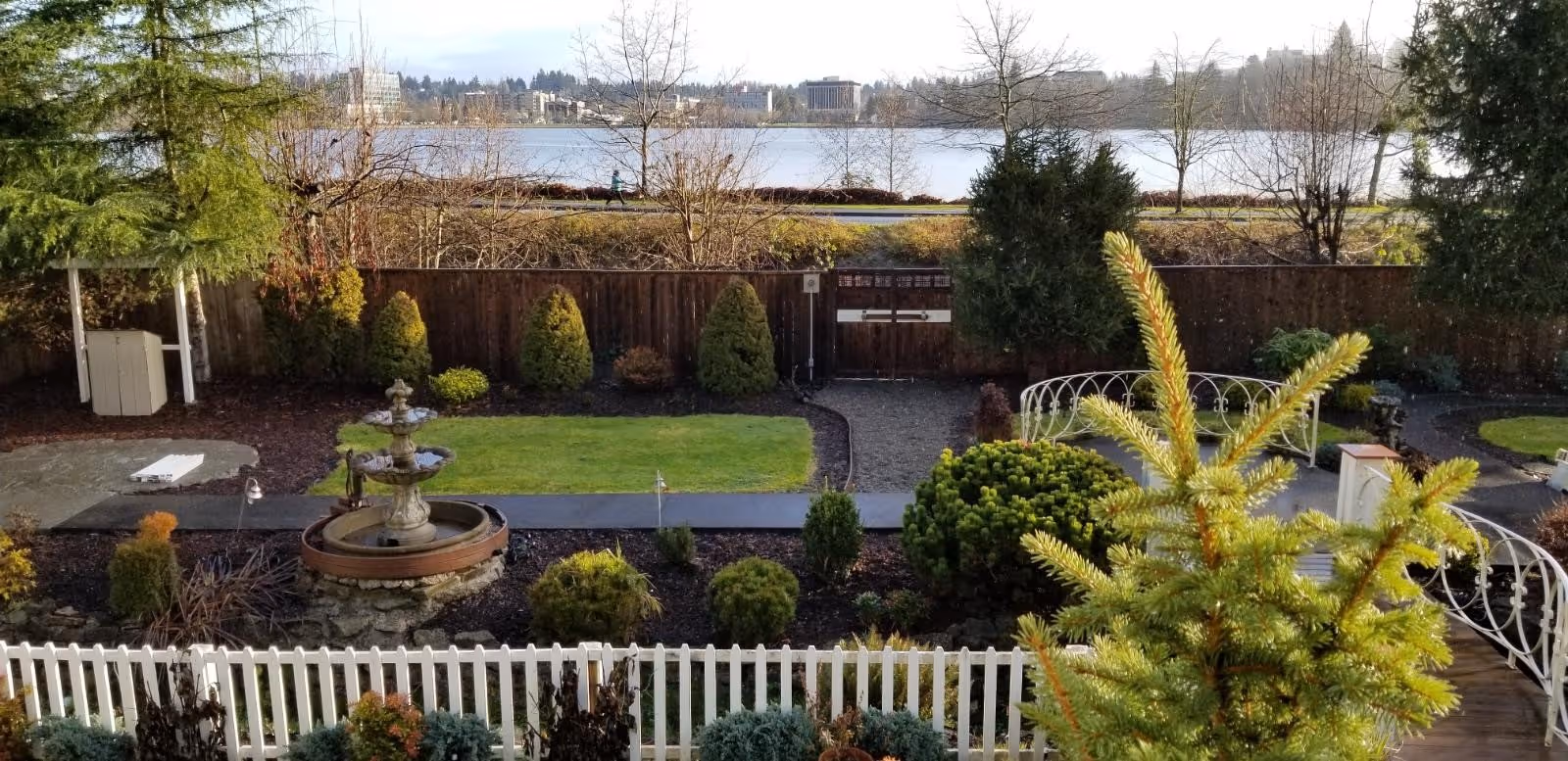 A landscaped outdoor garden area with a white picket fence in the foreground, a multi-tiered stone fountain, various shrubs and small trees, a gravel pathway, and a wooden fence in the background. Beyond the fence is a body of water with buildings visible on the opposite shore under a clear sky.