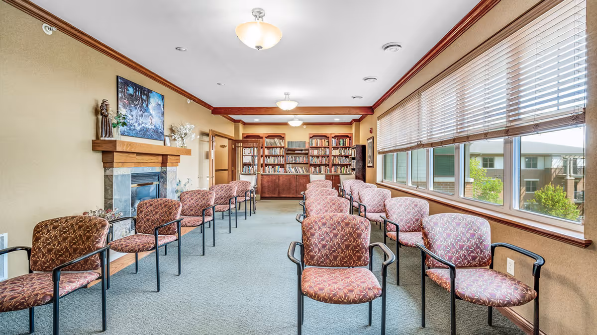 A well-lit room with rows of patterned chairs arranged facing a fireplace with a wooden mantel. The room has large windows with blinds on one side, letting in natural light. At the far end, there is a large bookshelf filled with books. The walls are beige with wooden trim, and the ceiling has recessed lighting and hanging light fixtures.