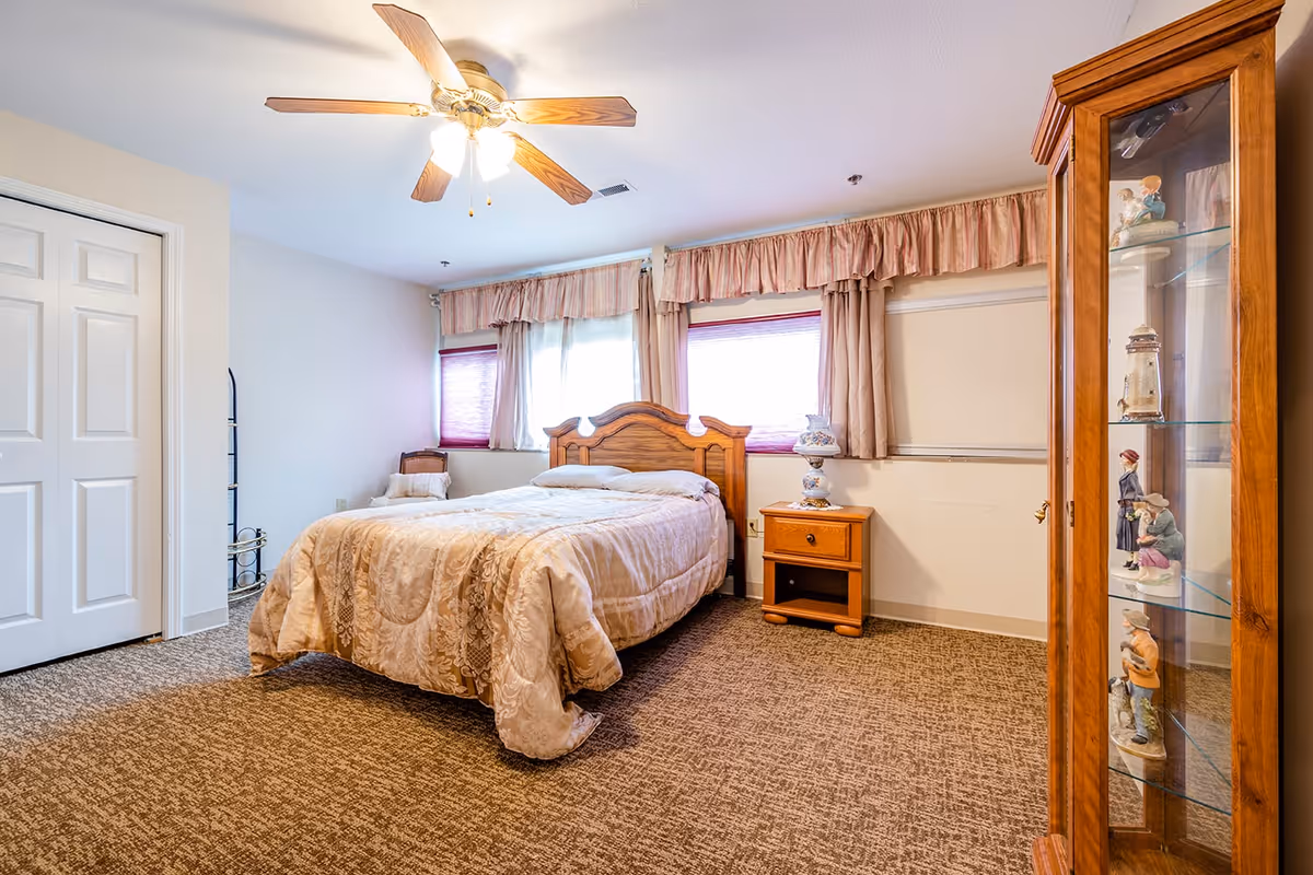 A cozy bedroom with a wooden bed frame and beige patterned bedding. The room has two windows with pink curtains and blinds, a wooden nightstand with a decorative lamp, a ceiling fan with lights, and a wooden display cabinet with figurines on glass shelves. The floor is carpeted in a brown pattern.
