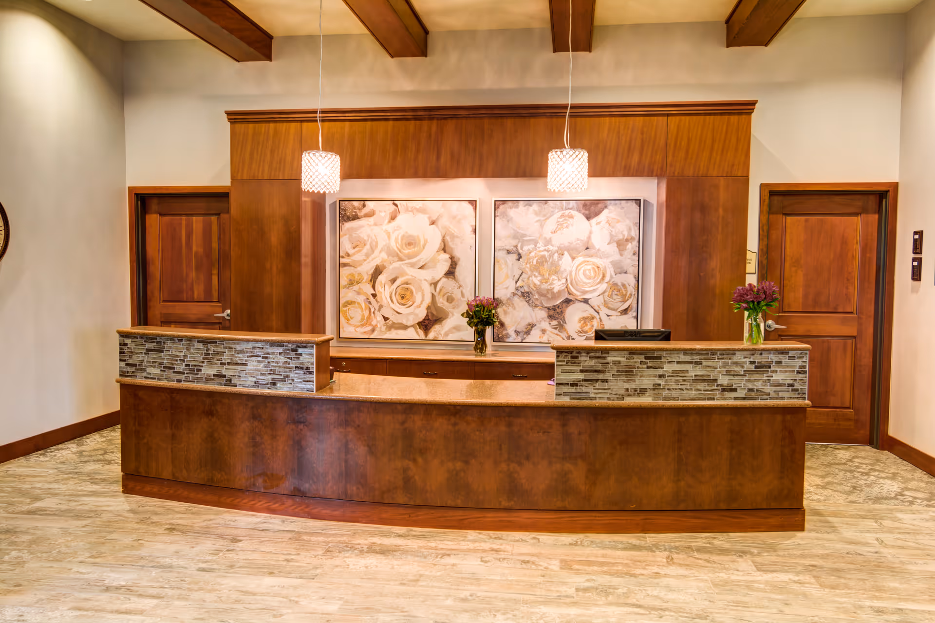 Reception desk area with a wooden counter featuring a stone tile accent, two pendant lights hanging from the ceiling, two wooden doors on either side, and two large floral paintings on the wall behind the desk. There are also two vases with flowers placed on the counter.