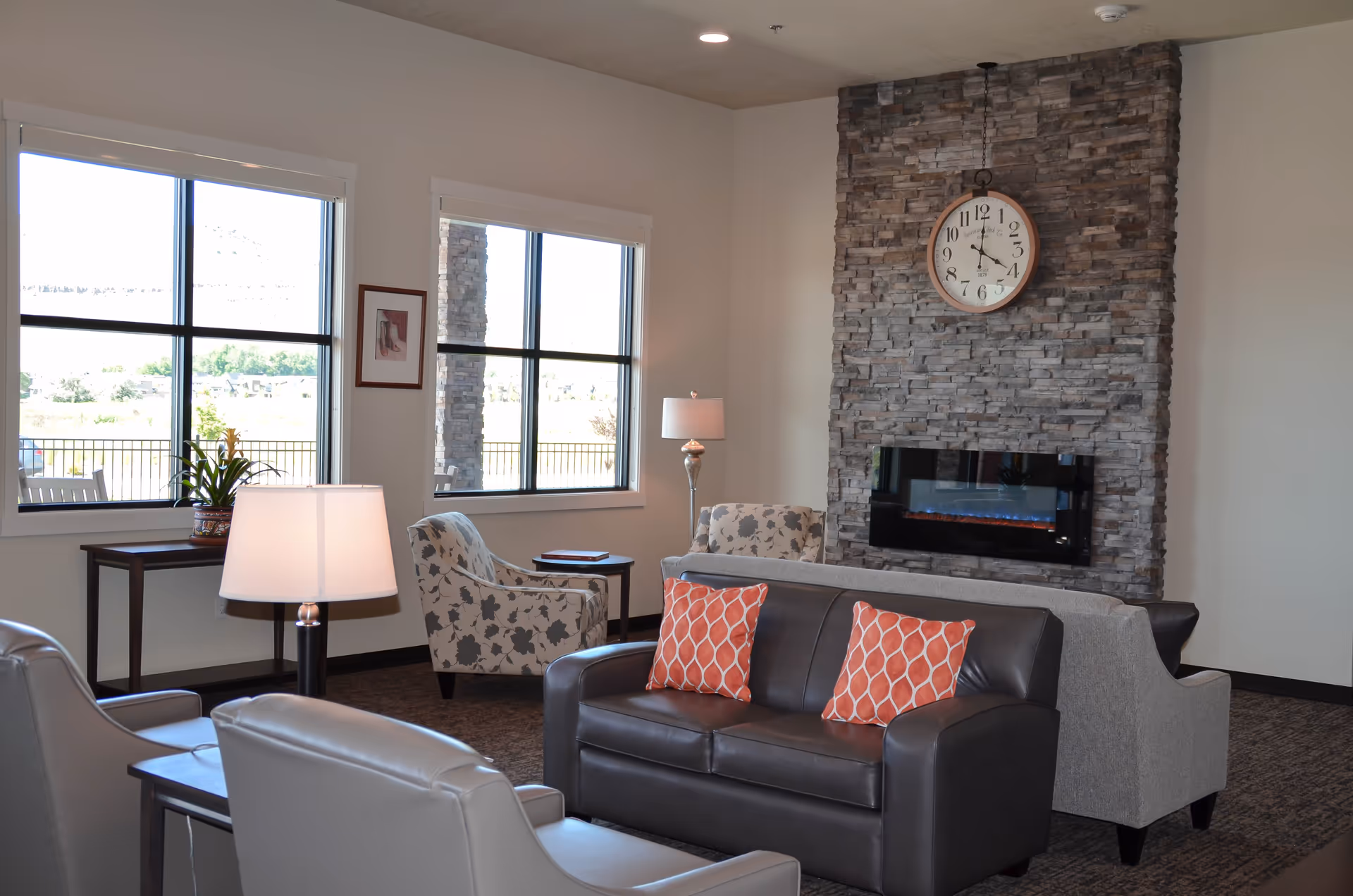 Bright common living room with sofas and armchairs facing a stone fireplace topped by a large clock, and large windows letting in daylight.