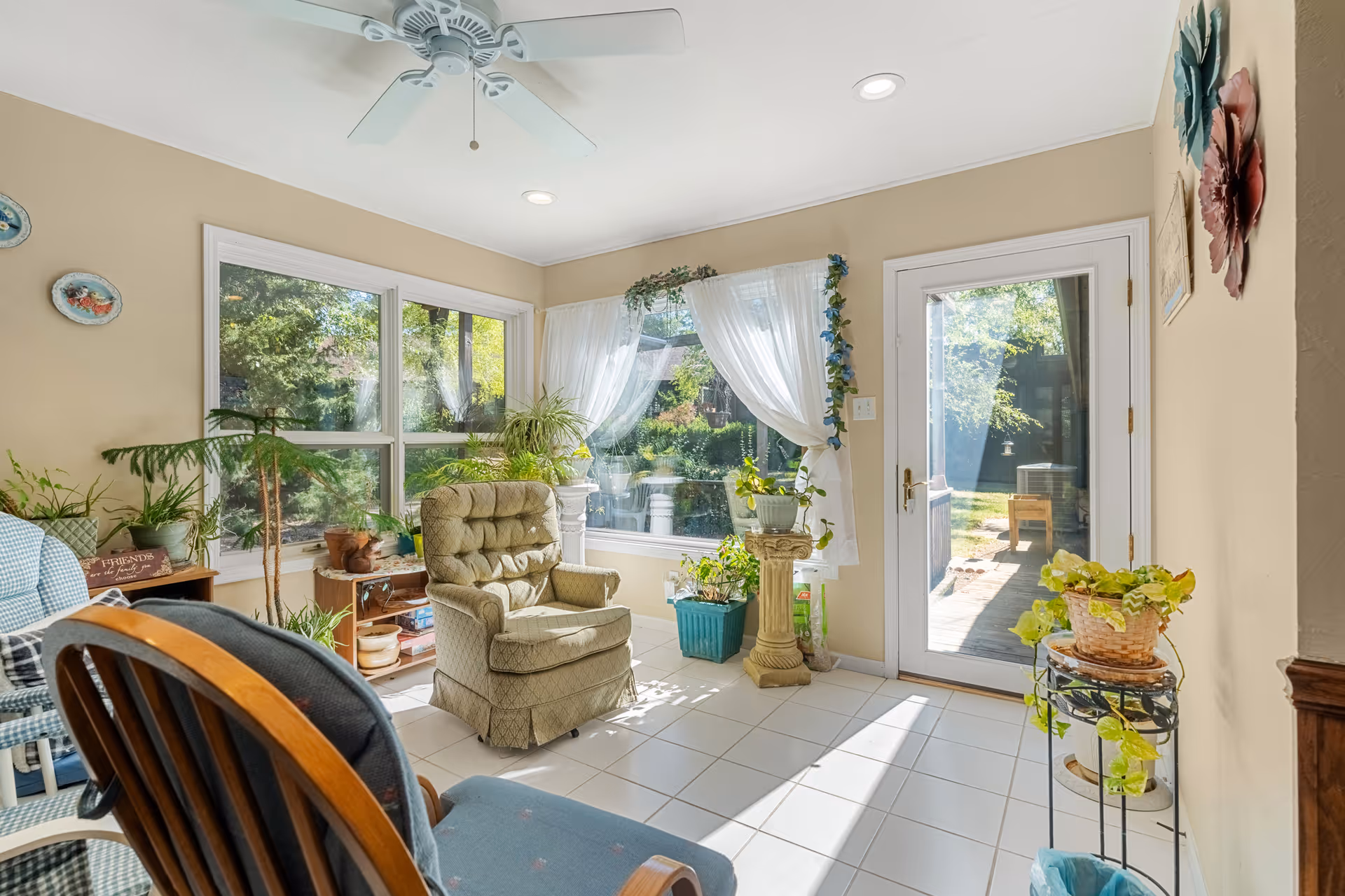 A bright sunroom with large windows and a glass door leading outside. The room contains several chairs, including a cushioned armchair and wooden rocking chair, and is decorated with various potted plants and light curtains. The walls are painted beige and the floor is tiled white.