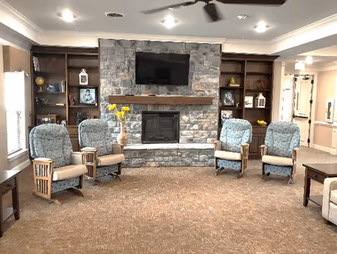 A cozy living room area with four cushioned armchairs arranged around a stone fireplace with a wooden mantel. Above the fireplace is a mounted flat-screen TV. Built-in wooden shelves flank the fireplace, decorated with framed photos, books, and decorative items. The room has beige walls, carpeted floor, and ceiling lights with a ceiling fan.