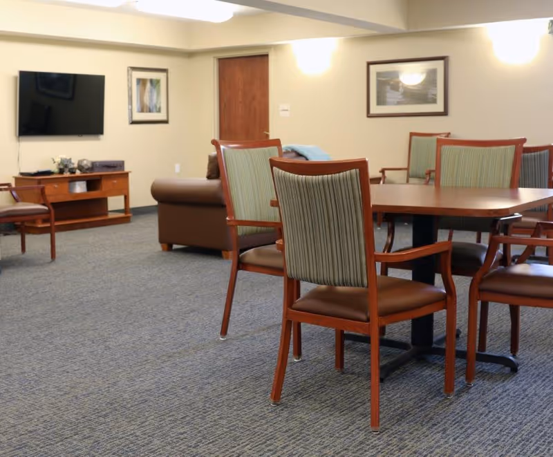A common area in a senior living facility with a round wooden table surrounded by six wooden chairs with green striped upholstery. In the background, there is a brown leather couch, a wooden TV stand with a flat-screen TV mounted on the wall, and framed artwork on the walls. The room has beige walls and carpeted flooring.