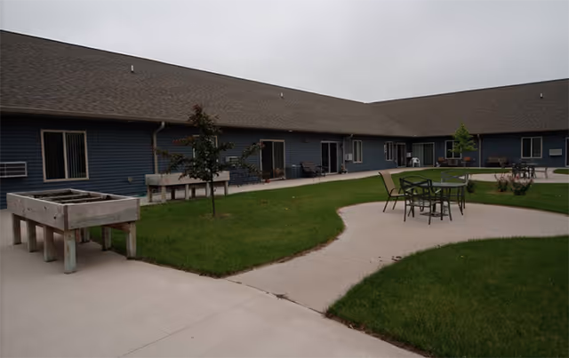 Courtyard of Prairie Place Assisted Living with a paved walkway, patio table and chairs, raised planters, lawn, and surrounding single-story building.