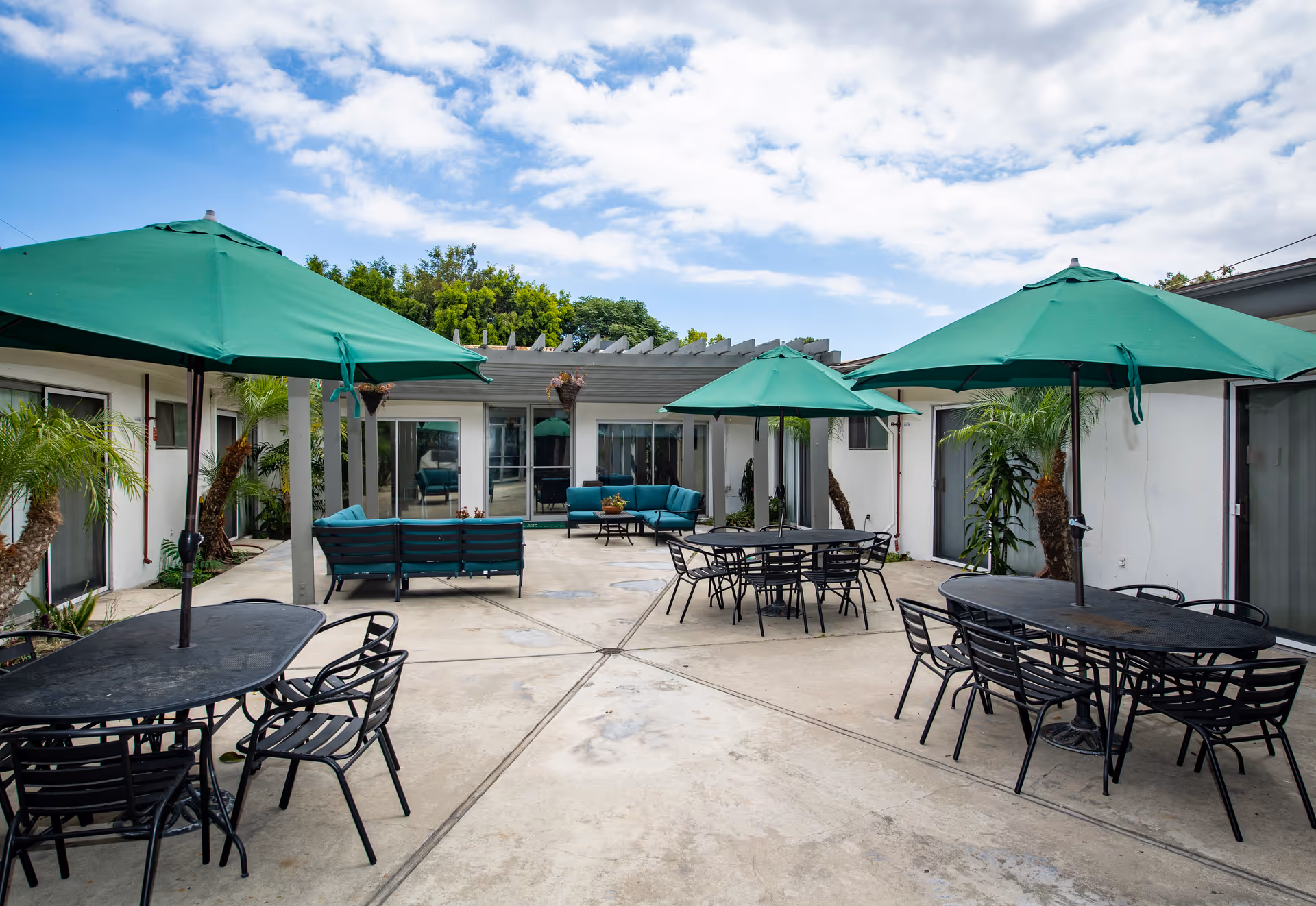 Outdoor courtyard area with several black metal tables and chairs, each shaded by large green umbrellas. There are also teal cushioned seating areas under a pergola attached to a white building. Palm trees and other greenery are planted around the perimeter, and the sky is partly cloudy.