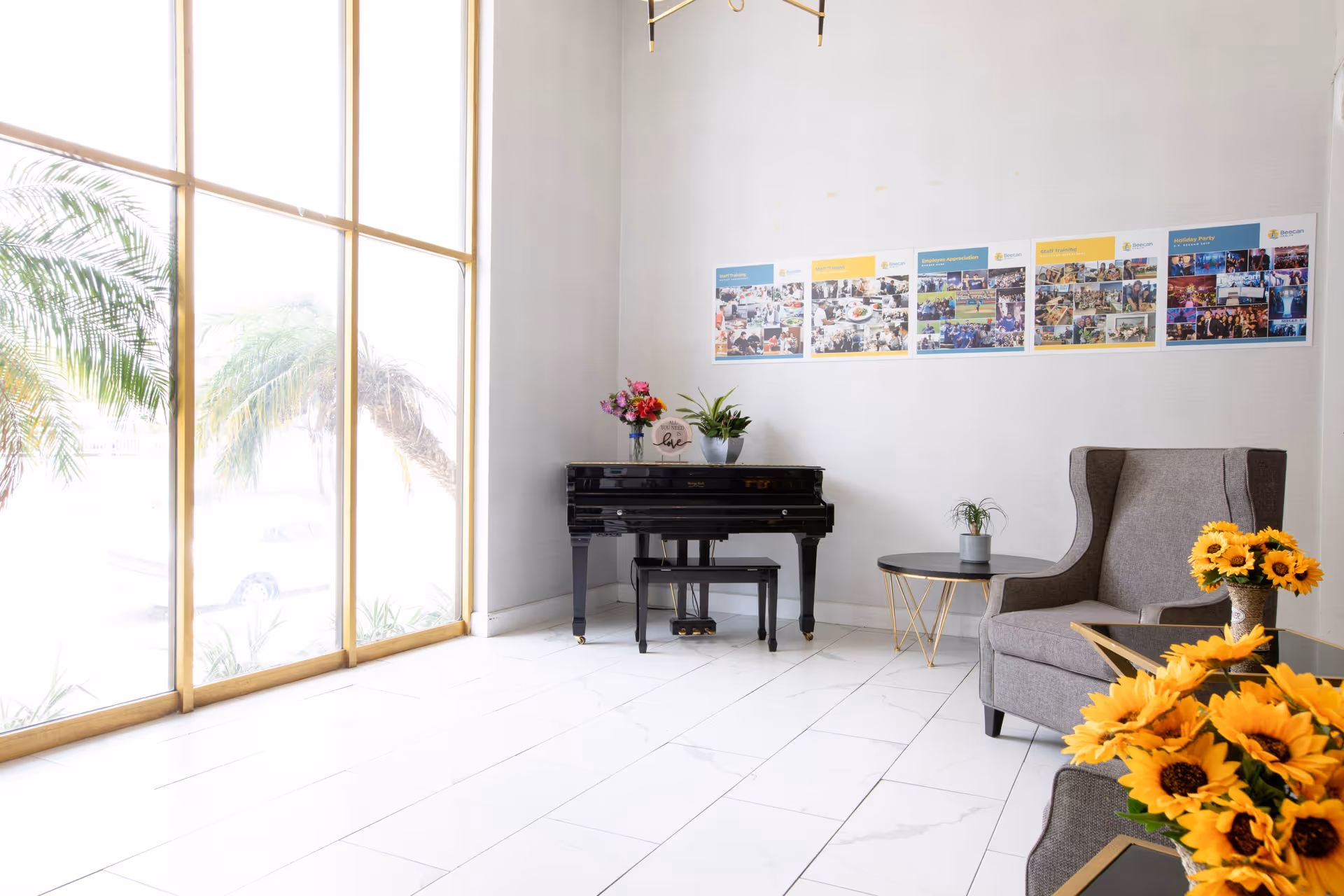 A bright and airy room with large floor-to-ceiling windows letting in natural light. The room features a black piano with a bench, decorated with a vase of flowers and a small plant on top. There is a comfortable gray armchair next to a round side table with a small plant. In the foreground, there is a table with a bouquet of sunflowers. The walls are white with a series of colorful posters or photo collages displayed on one wall.