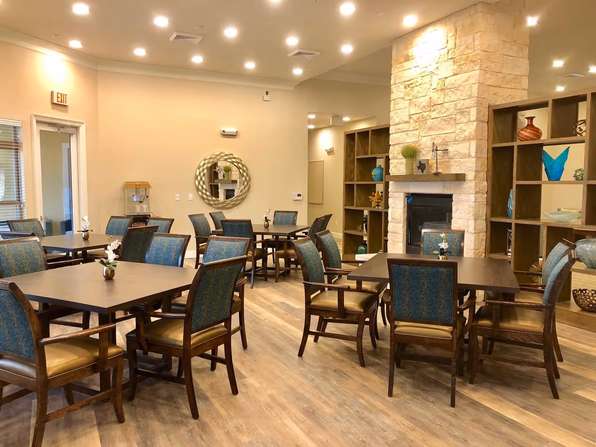 A well-lit dining area in a senior living facility with several wooden tables and chairs upholstered in blue and brown. The room features a stone fireplace, decorative shelving with vases and art pieces, a round wall mirror, and a popcorn machine near an exit door.