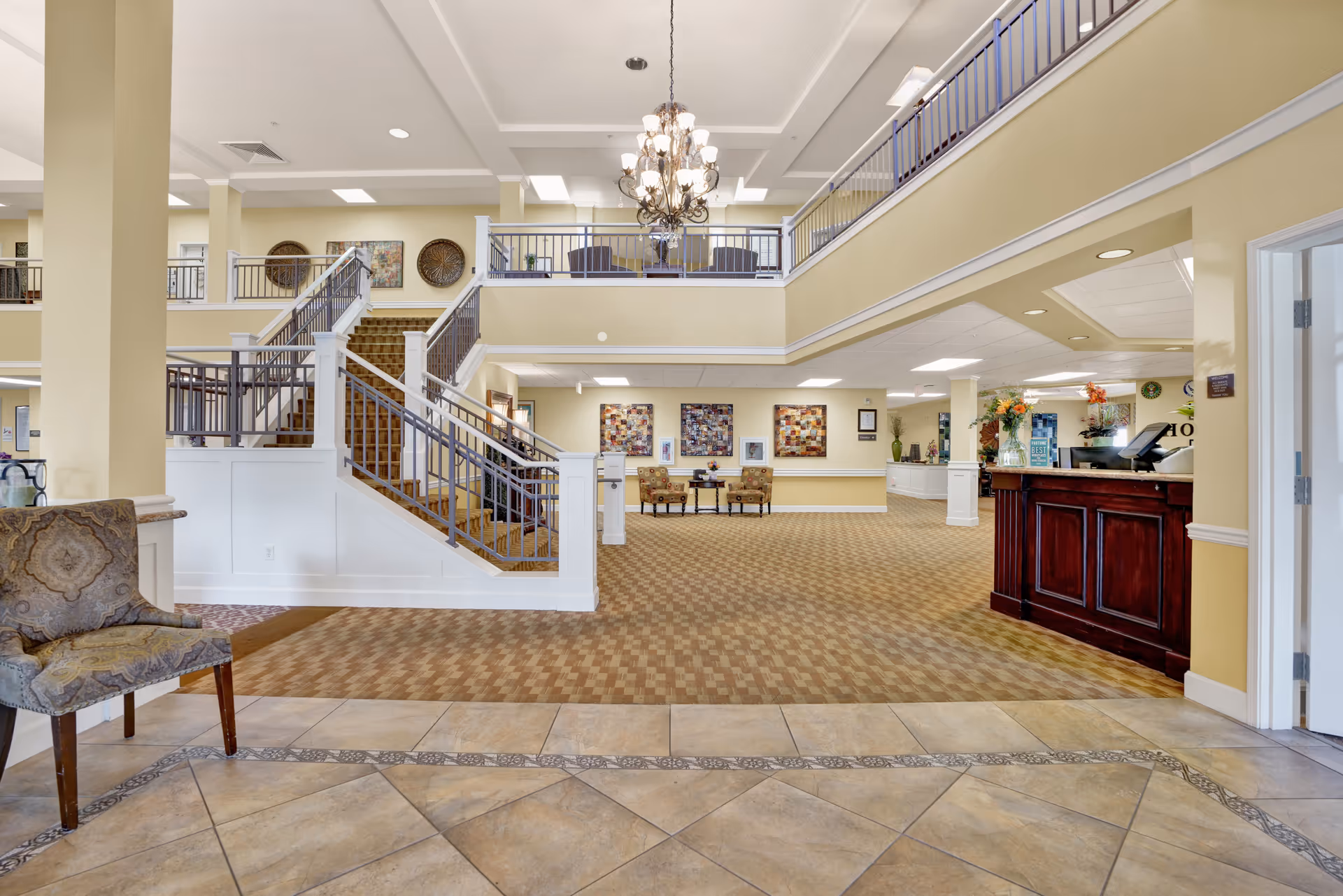 Spacious senior living facility lobby with a tiled floor entrance, patterned carpet, a wooden reception desk on the right, a staircase leading to an upper level with seating, and decorative wall art. The area is well-lit with a chandelier hanging from the ceiling.