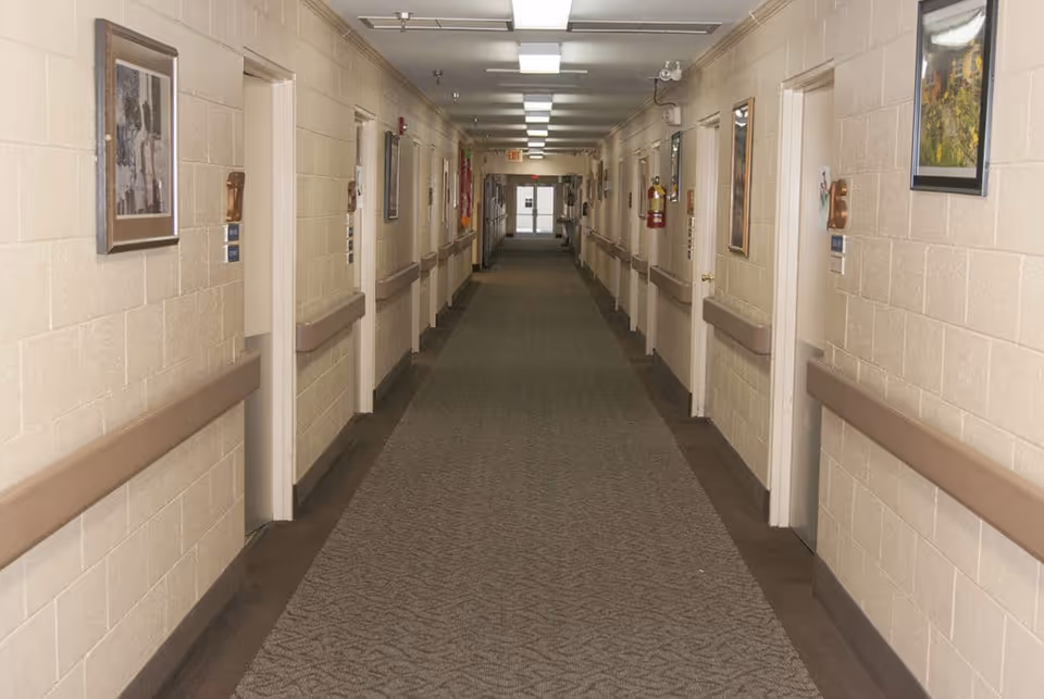 Long indoor hallway in a personal care home with beige walls, carpeted floor, handrails on both sides, multiple closed doors, framed pictures on the walls, and ceiling lights.