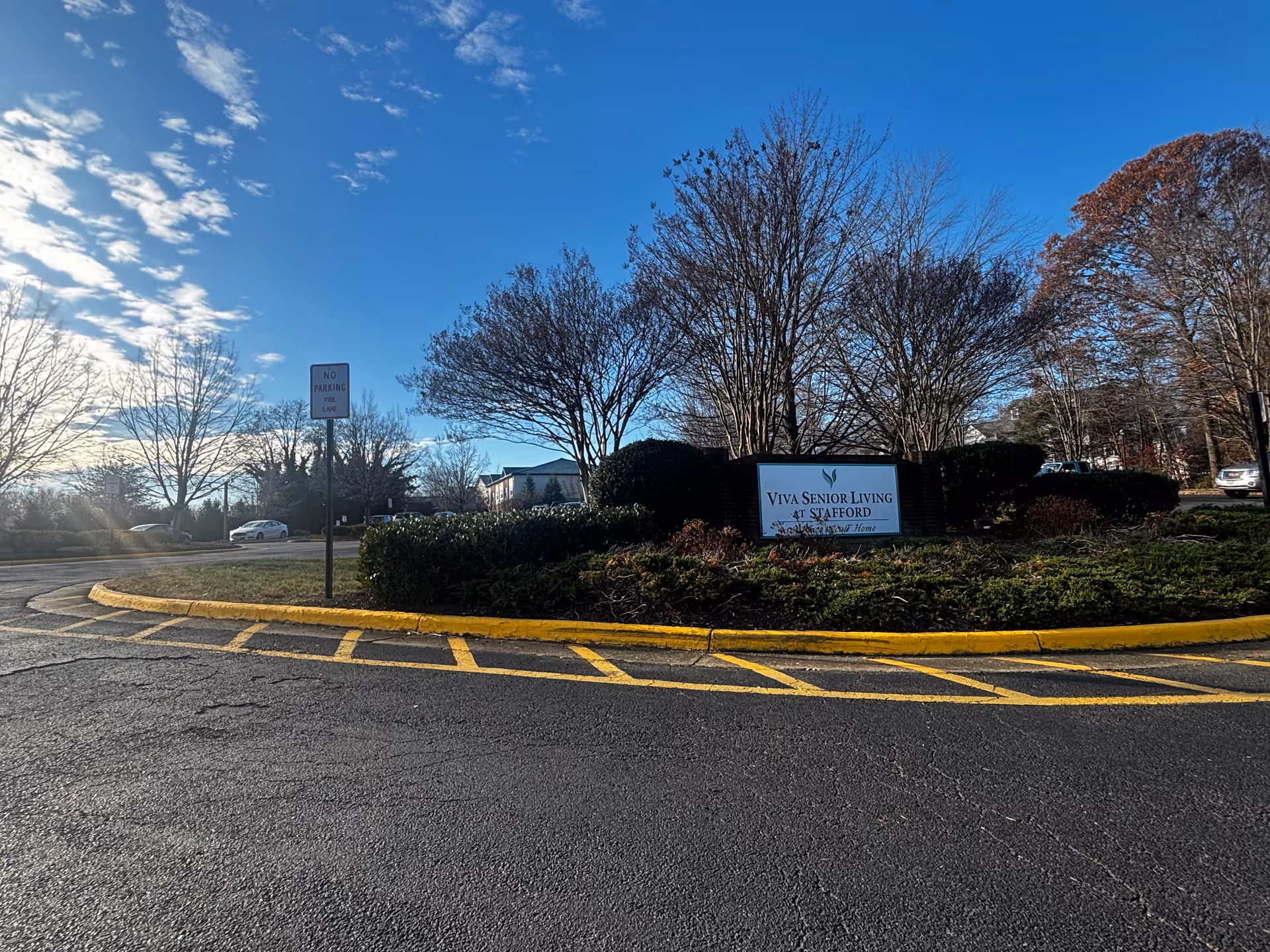 Outdoor view of the entrance area to a senior living facility with a sign that reads 'Viva Senior Living at Stafford' surrounded by bushes and trees under a blue sky with some clouds.