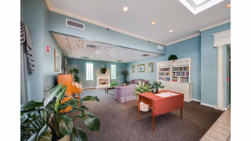 A cozy common area in a senior living facility with light blue walls, a carpeted floor, and a skylight. The room features a striped sofa, a wooden coffee table, a white bookshelf filled with books, several framed pictures on the walls, a piano, and various potted plants. There is also a small table with a red tablecloth near the entrance.