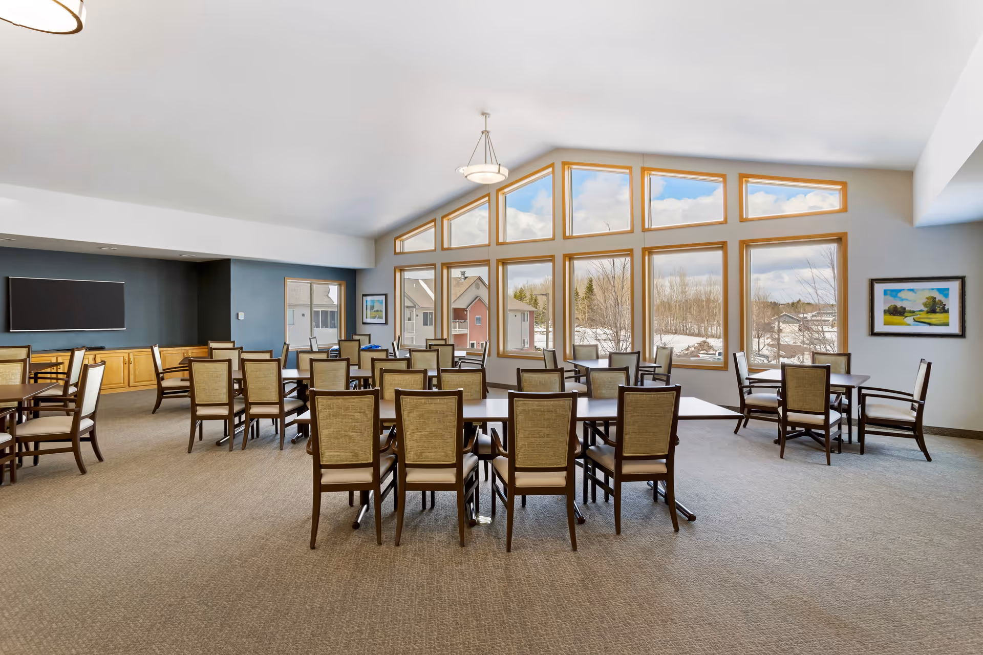 A spacious dining room with multiple tables and chairs arranged neatly. Large windows with wooden frames allow natural light to fill the room and offer a view of houses and trees outside. The room has a high ceiling with a hanging light fixture and a wall-mounted flat screen TV on a dark accent wall.