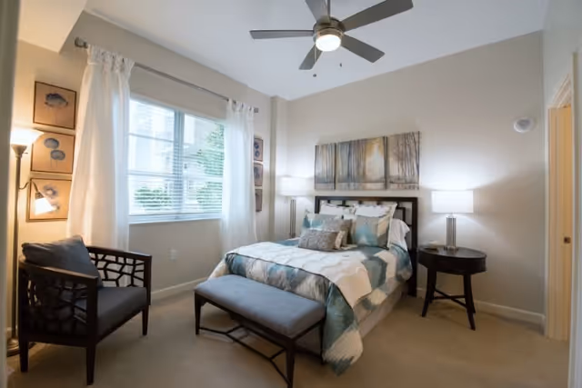 Sunlit furnished bedroom with a bed, bench, armchair, nightstands and a ceiling fan.