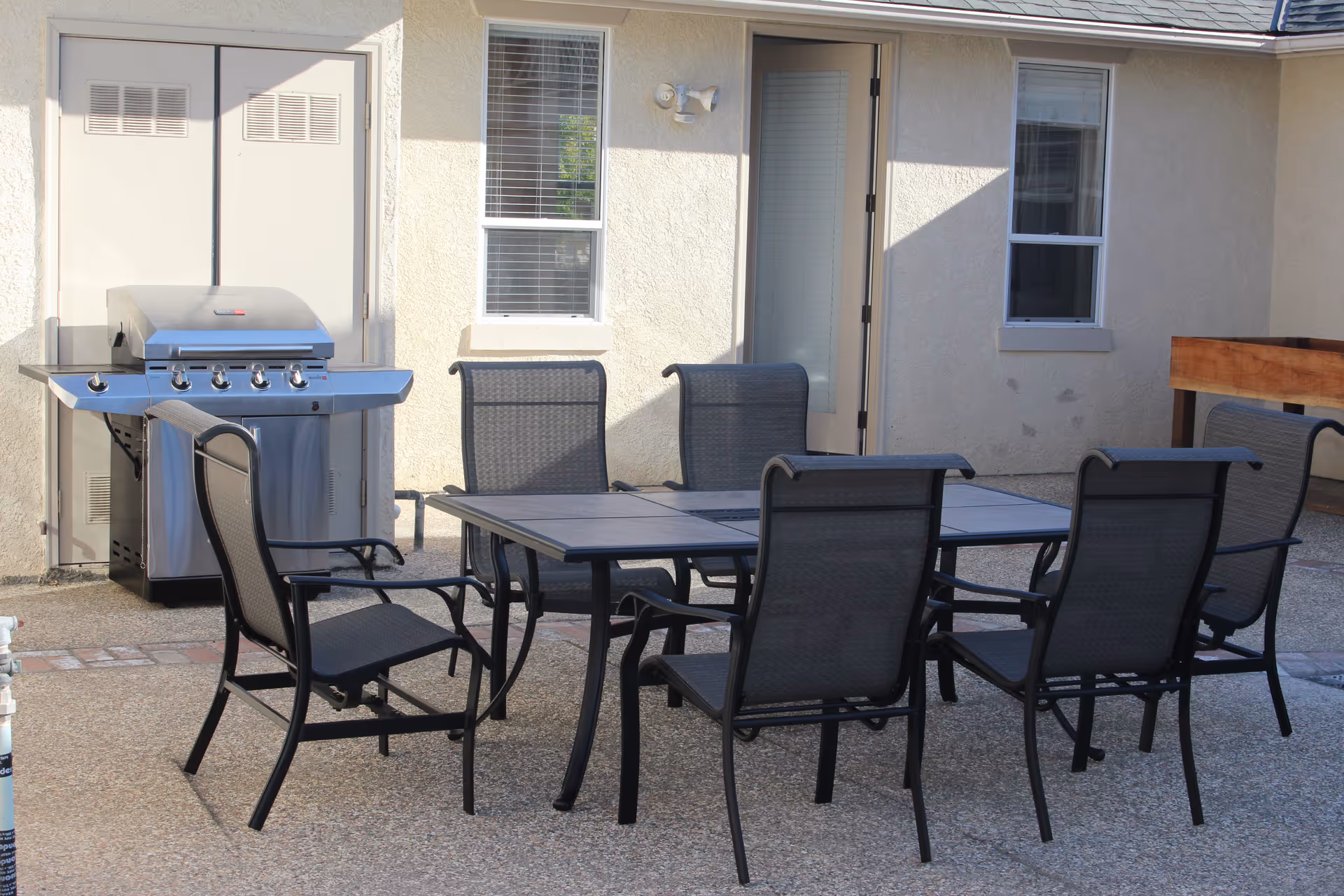 Patio area with a rectangular outdoor dining table surrounded by six black mesh chairs and a stainless-steel grill against a beige building wall with a door and windows.