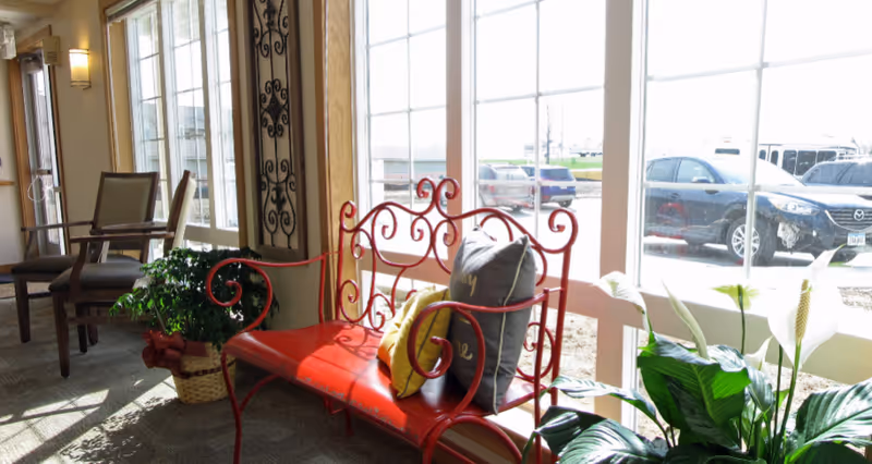 A bright interior seating area with a decorative red metal bench adorned with two pillows, one yellow and one gray. Next to the bench is a green potted plant in a basket. Large windows behind the bench let in natural light and show parked cars outside. Additional chairs and wall decorations are visible in the background.