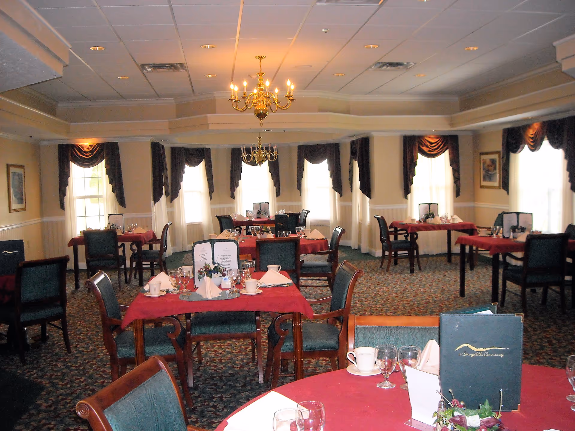 A dining room with multiple tables covered in red tablecloths, set with glassware, cups, napkins, and menus. The room has large windows with dark curtains, chandeliers hanging from the ceiling, and patterned carpet flooring.
