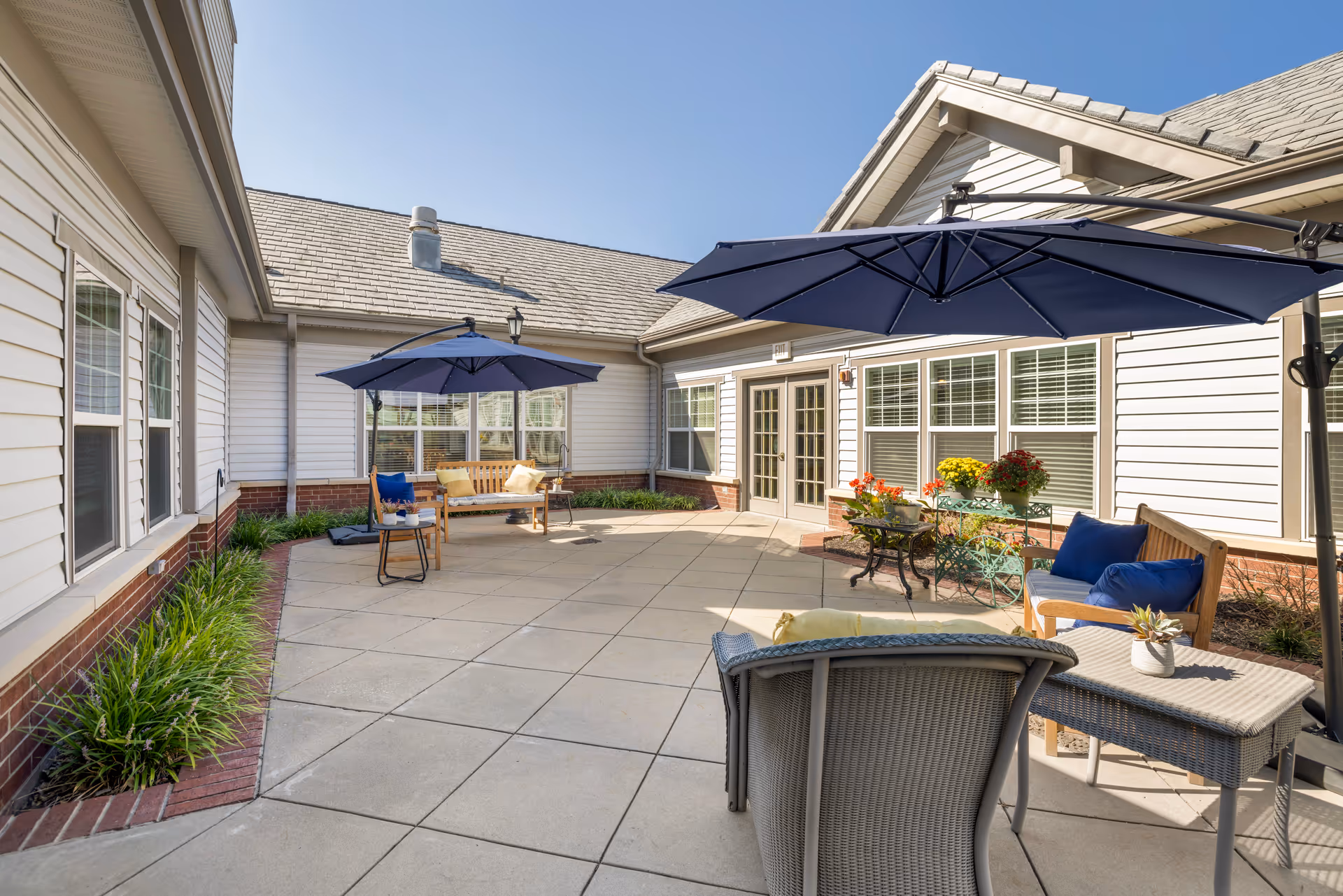 Sunny enclosed courtyard patio with tiled flooring, blue umbrellas, outdoor seating and potted flowers outside a single-story building.