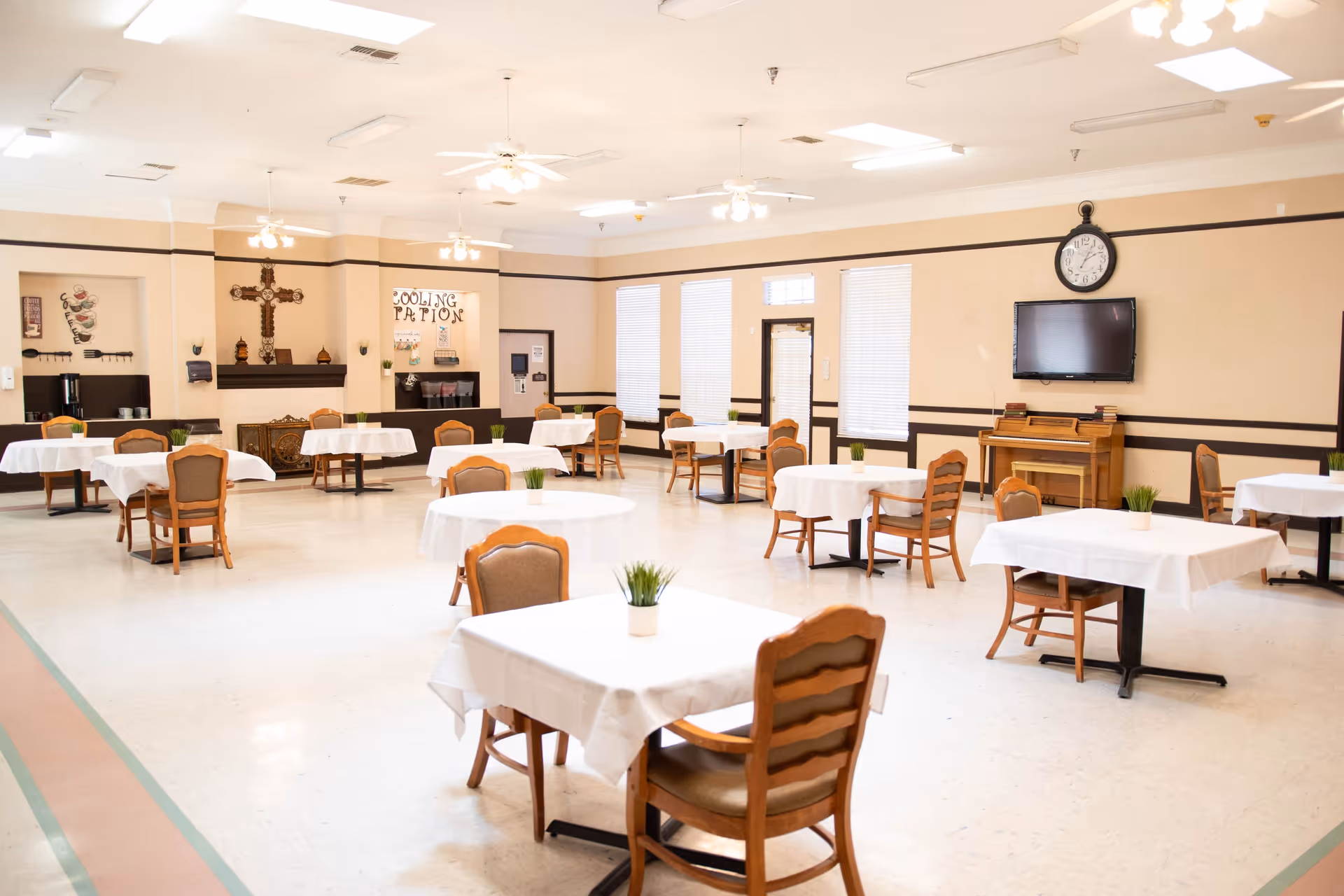 A spacious dining room with multiple round and square tables covered with white tablecloths, each table having a small potted plant centerpiece. Wooden chairs surround the tables. The room has beige walls with dark trim, ceiling fans with lights, a wall-mounted TV above a piano, and large windows with blinds letting in natural light.