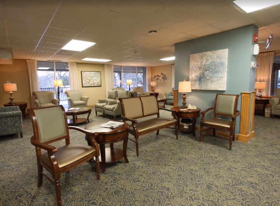 A spacious senior living lounge with upholstered chairs, wooden tables, lamps, and wall art.