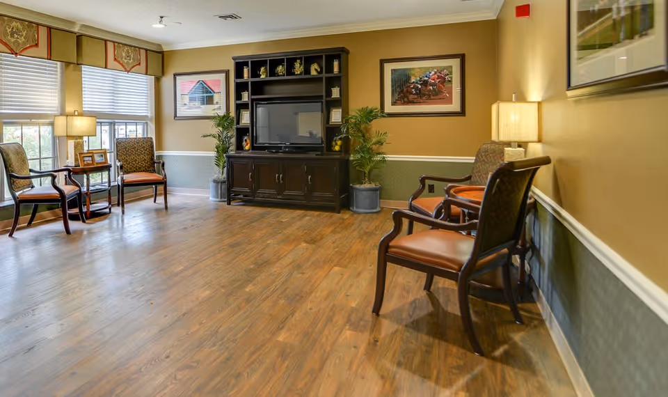 A bright common sitting area with upholstered chairs, side tables, lamps, potted plants and an entertainment cabinet on wood floors.