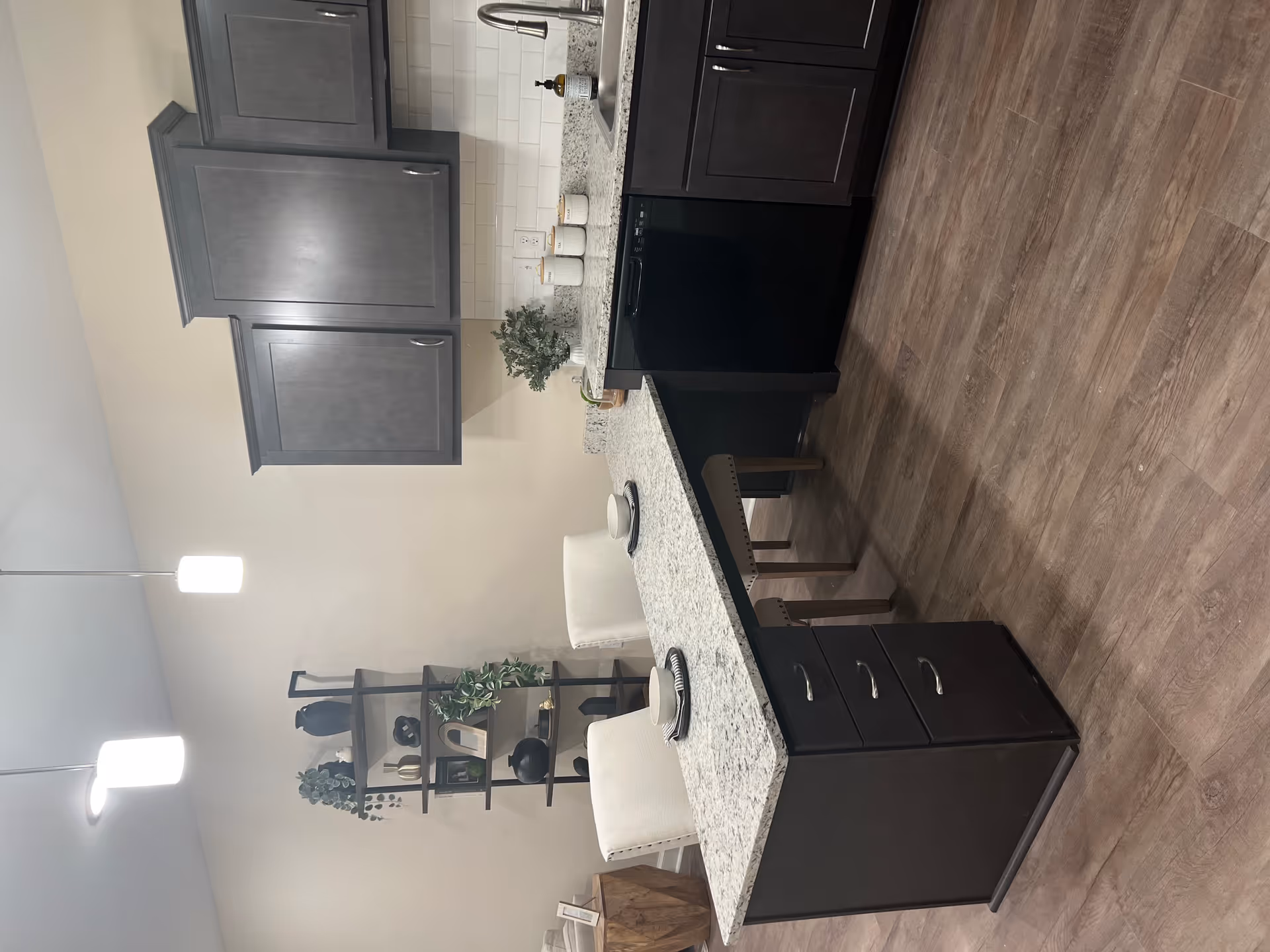 Modern kitchen area with dark wood cabinets, granite countertops, a built-in dishwasher, and a sink. A peninsula with drawers extends into the room with two white cushioned chairs set with bowls and plates. In the background, a tall black metal shelf holds decorative items and plants. The floor is wood-style laminate and two pendant lights hang from the ceiling.