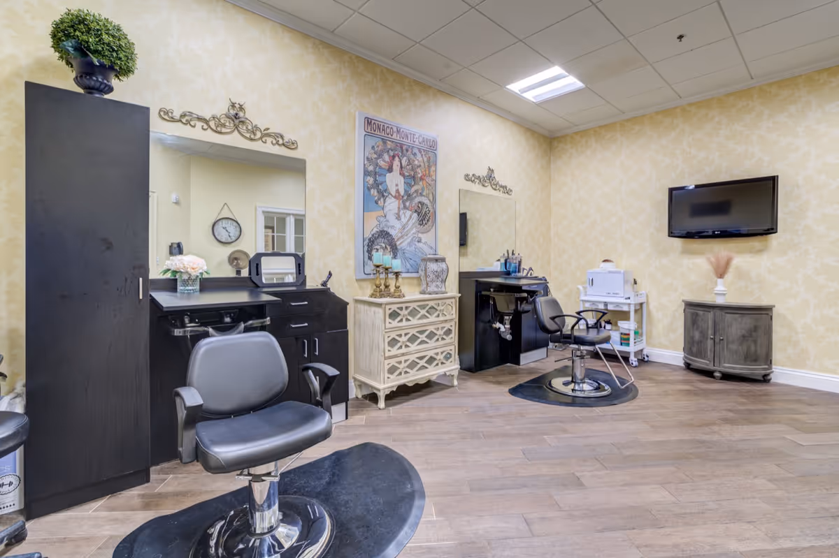 Interior view of a salon area with two black salon chairs in front of mirrors and black cabinetry. There is a decorative cabinet with candles and a framed Monaco-Monte Carlo poster on the wall. A flat-screen TV is mounted on the wall, and the room has wood flooring and yellow patterned wallpaper.