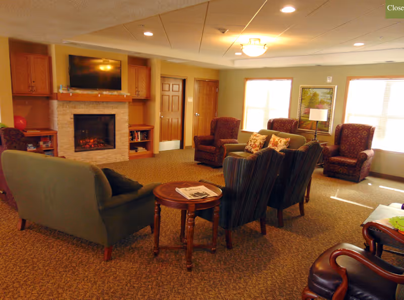 A cozy living room area with a green sofa, two striped armchairs, and two patterned armchairs arranged around a small wooden round table with a newspaper on it. There is a fireplace with a TV mounted above it, wooden cabinets on either side, and two wooden doors in the background. The room is carpeted and has windows letting in natural light, with a framed painting and a floor lamp near the windows.