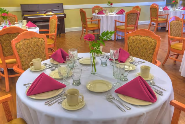 A dining room set up with round tables covered in white tablecloths, each table set with beige plates, cups, silverware, clear glasses, and folded magenta napkins. A small vase with a pink flower and green leaves is placed at the center of each table. Wooden chairs with patterned upholstery surround the tables. A piano is visible against the far wall.