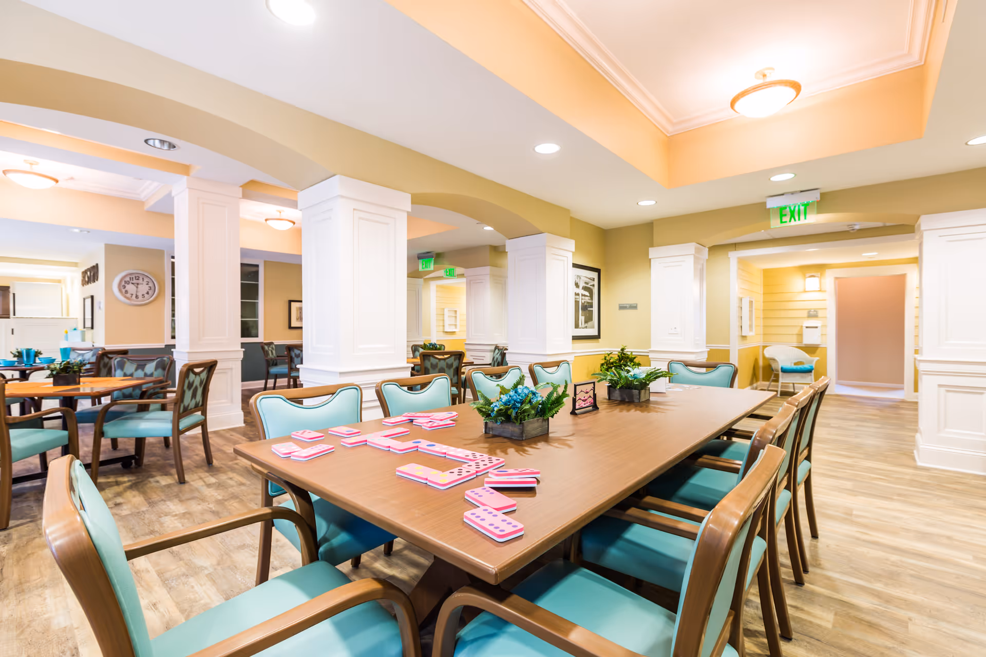 A bright and spacious common area in a senior living facility featuring a large wooden table with turquoise cushioned chairs around it. The table has dominoes and small potted plants on it. The room has wooden flooring, white pillars, and warm yellow walls with recessed lighting and ceiling fixtures. Additional seating and tables are visible in the background along with a clock on the wall.