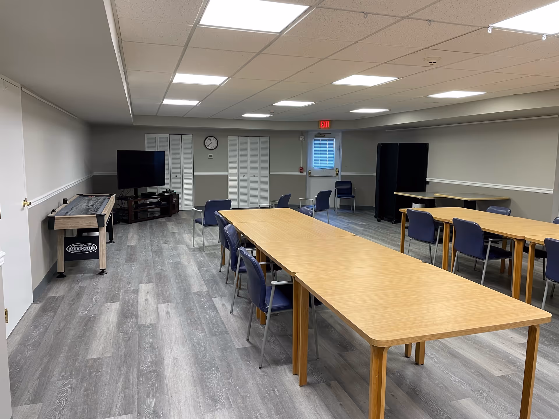 A spacious room with light gray walls and wood-patterned flooring, featuring several wooden tables arranged in rows with blue chairs around them. At the far end, there is a shuffleboard table, a flat-screen TV on a stand, a clock on the wall, and a door with an exit sign above it. The ceiling has recessed lighting panels.