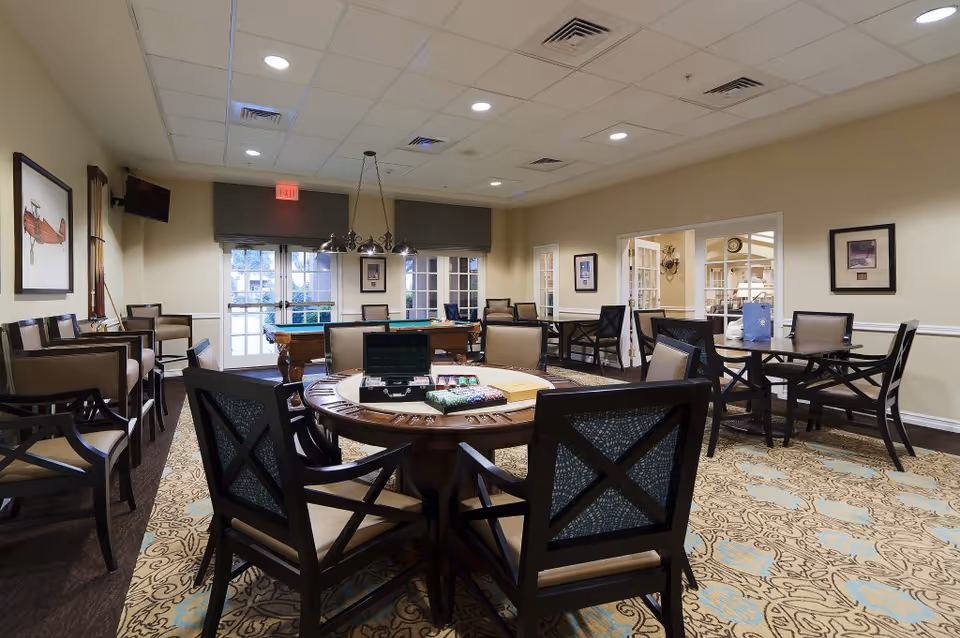 A well-lit common room with multiple tables and chairs arranged for social activities. In the center, a round table has a poker set and chips, while in the background there is a pool table near large windows and glass doors. The walls are decorated with framed pictures, and the room has a patterned carpet and recessed ceiling lights.