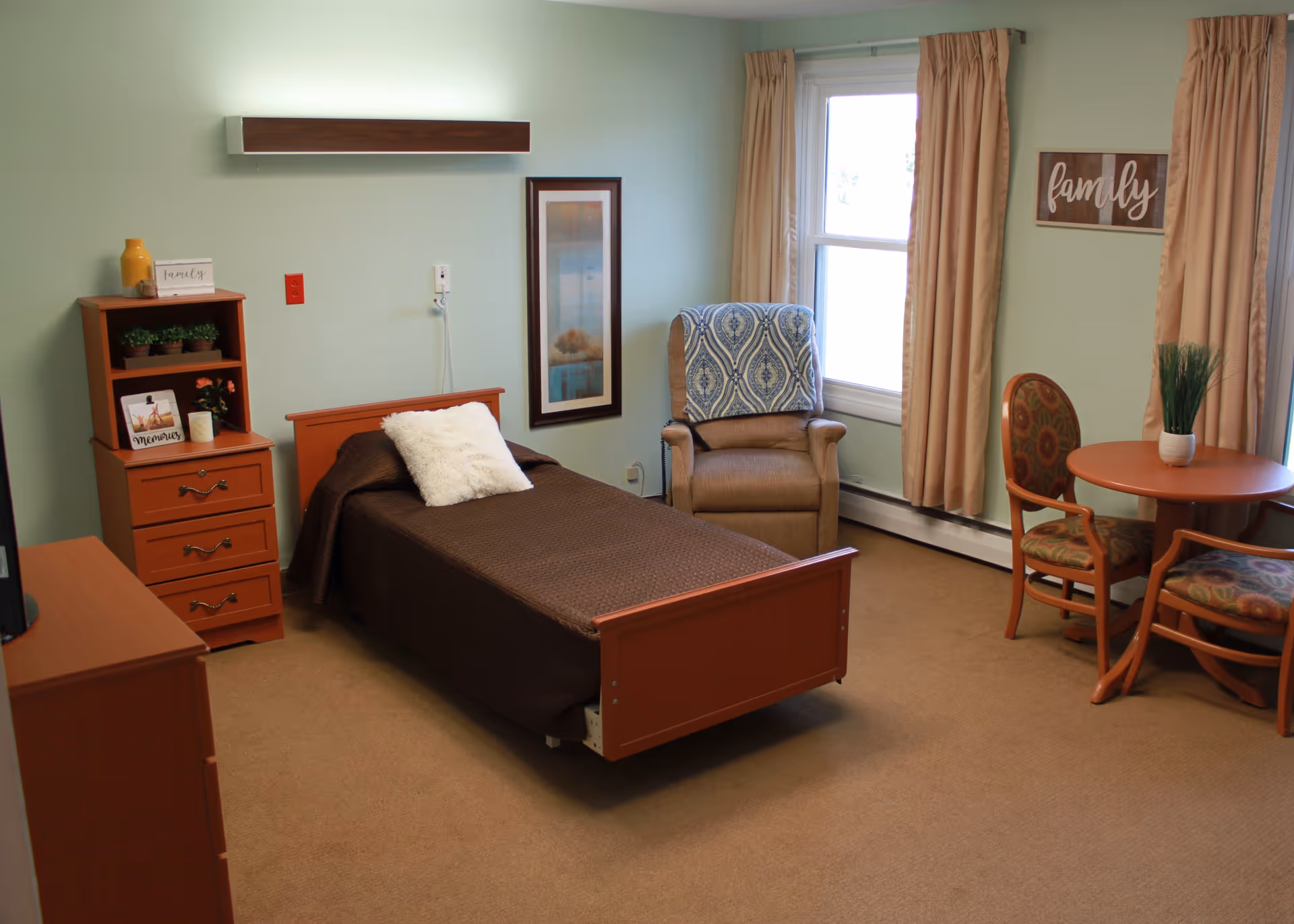 A cozy bedroom in a senior living facility with a single bed covered in a brown blanket and a white pillow. Next to the bed is a wooden dresser with decorative items including a yellow vase and small plants. A comfortable armchair with a patterned cover is placed near a window with beige curtains. A small round table with two upholstered chairs is also in the room. The walls are painted light green and decorated with framed artwork and a sign that says 'family'.