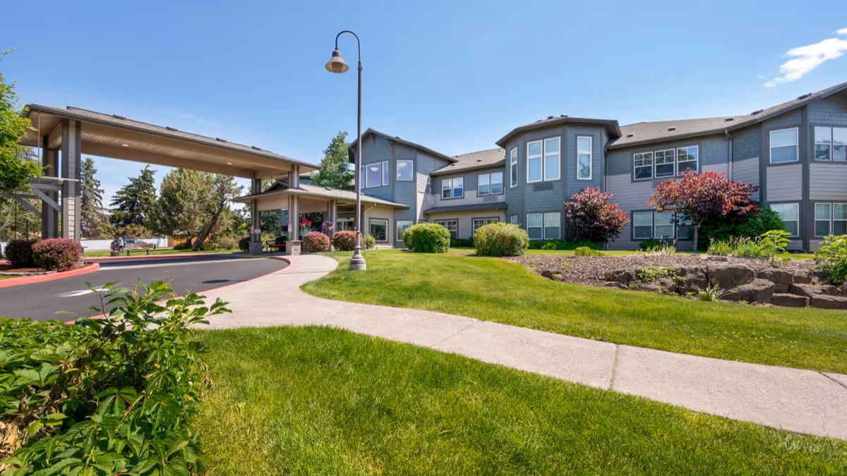 Front exterior of a senior living building with a covered entrance, landscaped lawn, and walkway.
