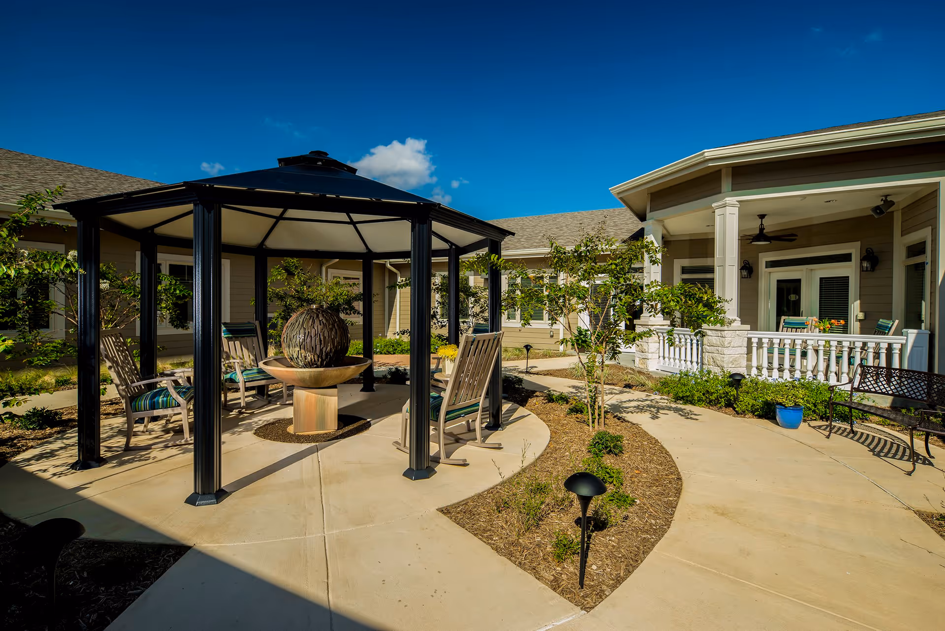 Outdoor courtyard area at Chapters Living of New Braunfels featuring a black metal gazebo with seating around a decorative water fountain, surrounded by landscaped plants and trees, with a building porch and seating area in the background under a clear blue sky.